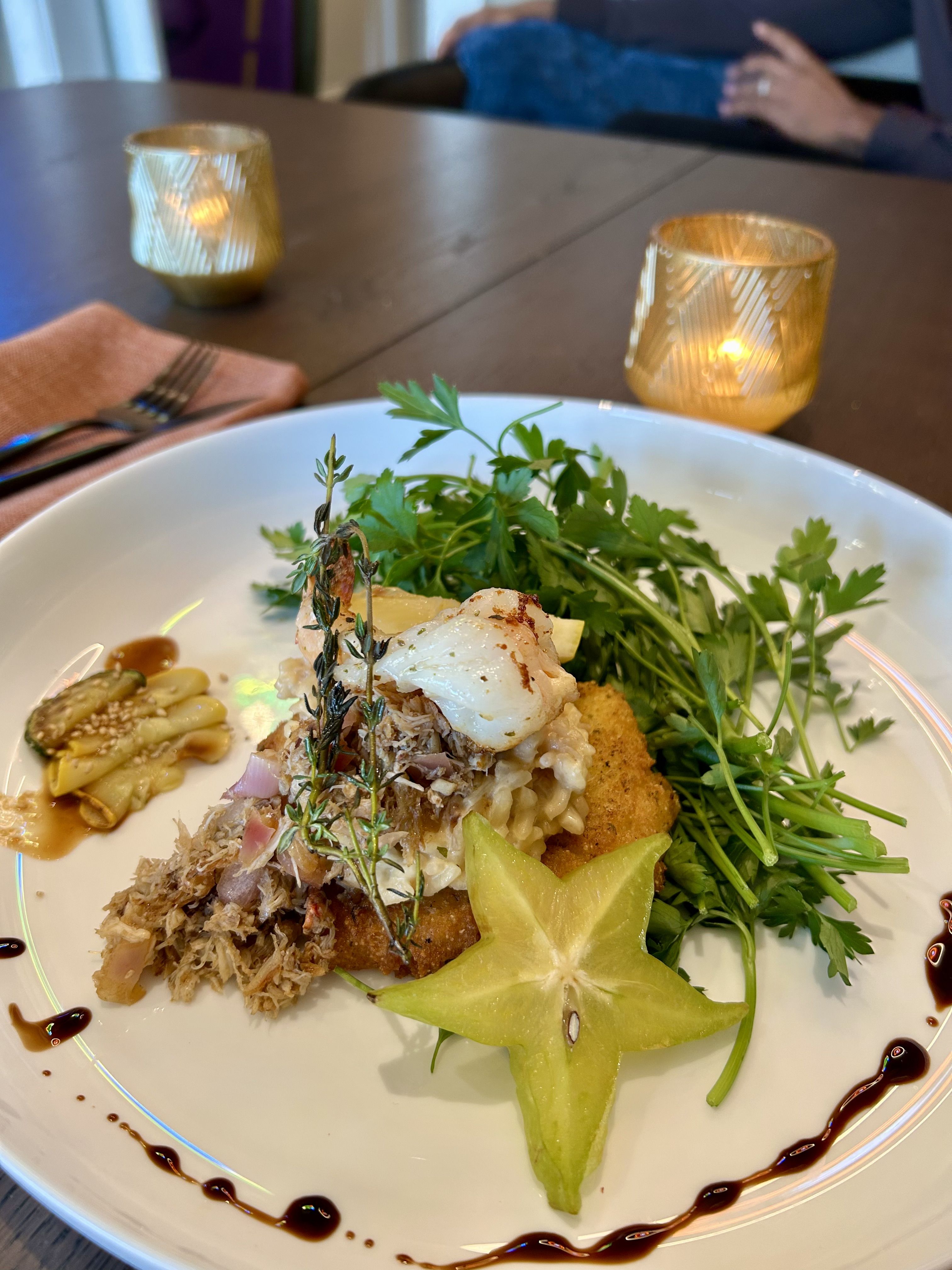 Gourmet dish with grilled seafood, herbs, shredded meat, risotto, starfruit slice, and sauce drops on a white plate, set on a wooden table with lit candle holders nearby.