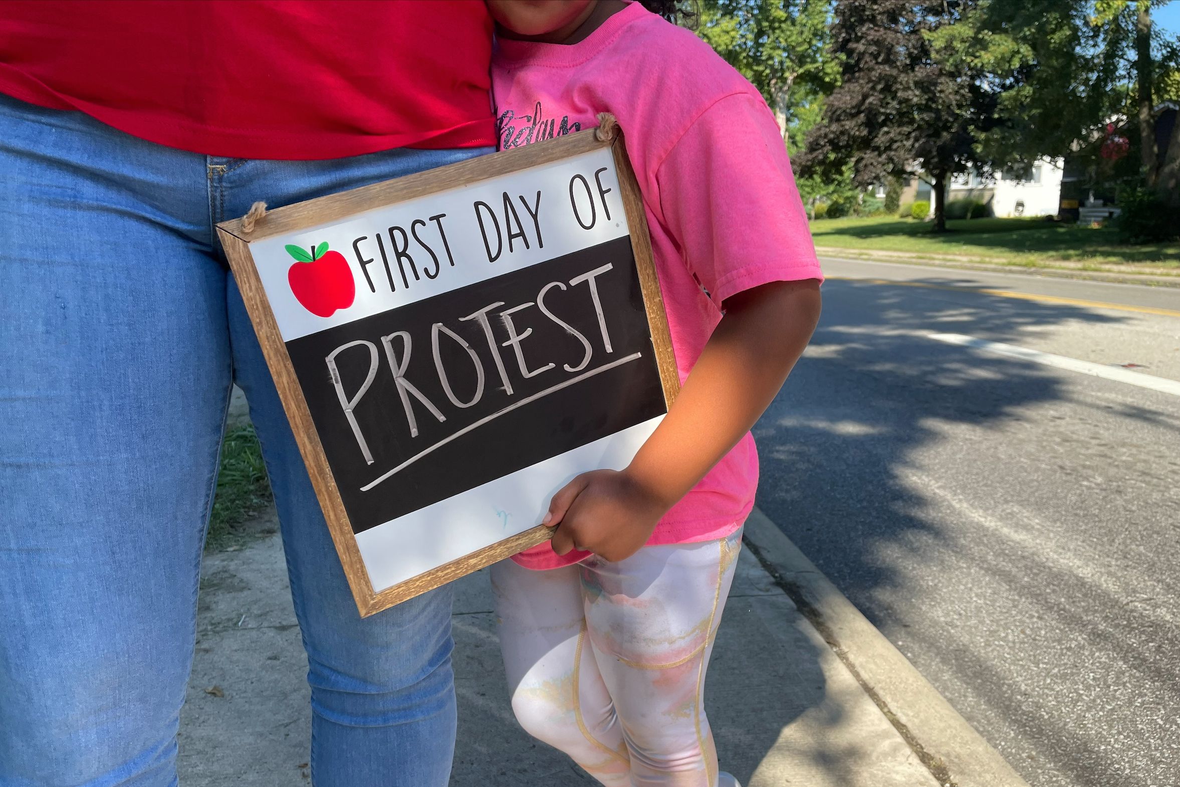 A young girl holds a sign next to her mother reading "First Day of Protest."