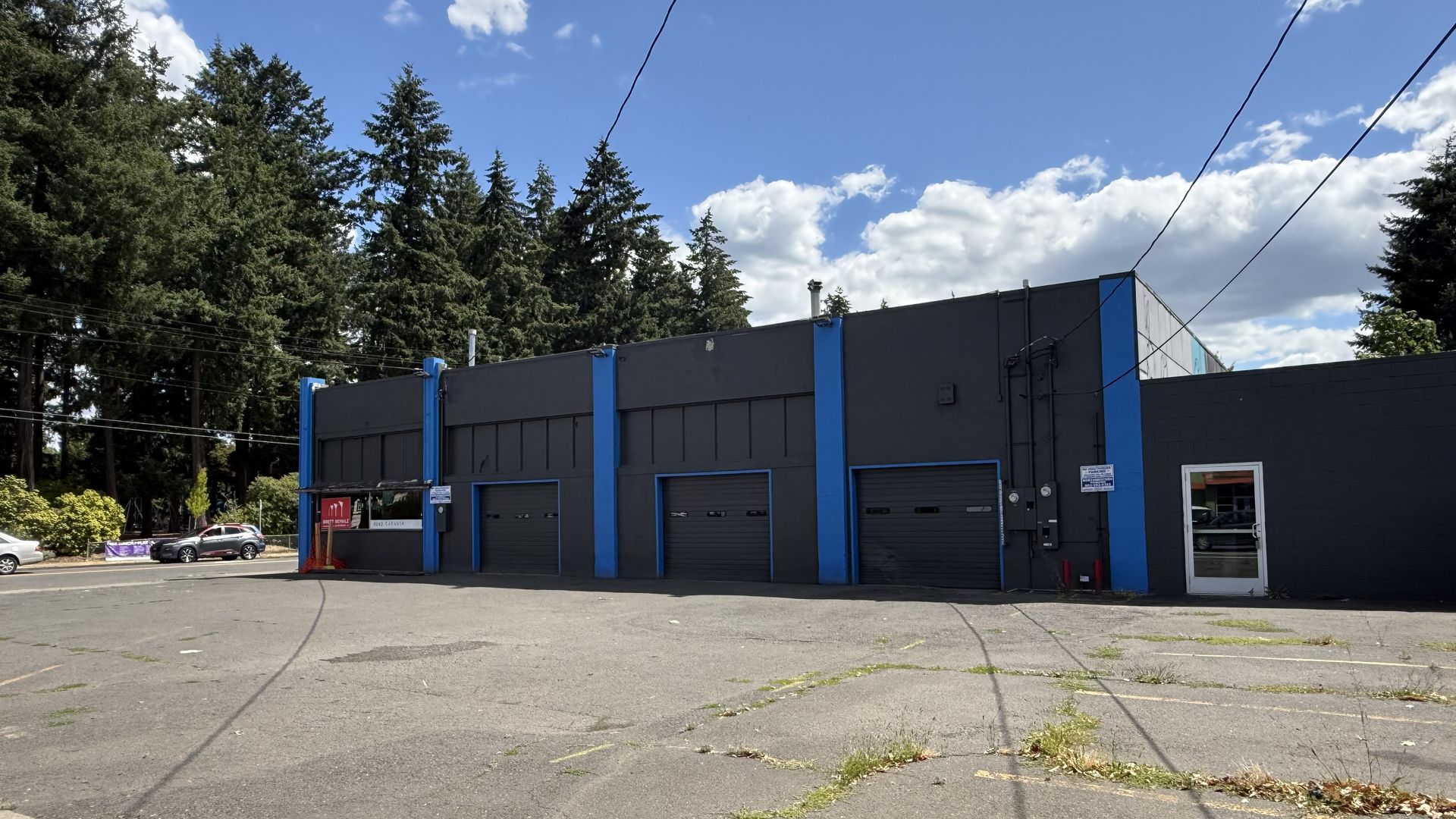 Industrial gray building with blue vertical accents and garage doors sits in a paved lot with cars on the left; tall evergreen trees, blue sky and white clouds behind, power lines overhead.