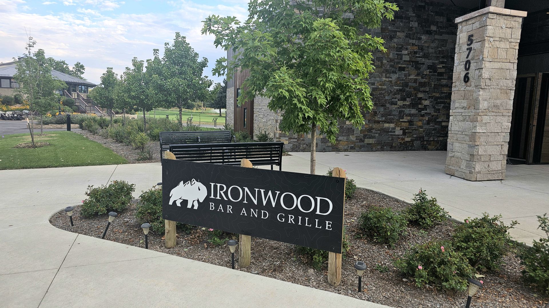 Outdoor view of Ironwood Bar and Grille: a black sign with a white bear logo in front of green trees, a stone building pillar, benches, and landscaped shrubs under a blue sky.
