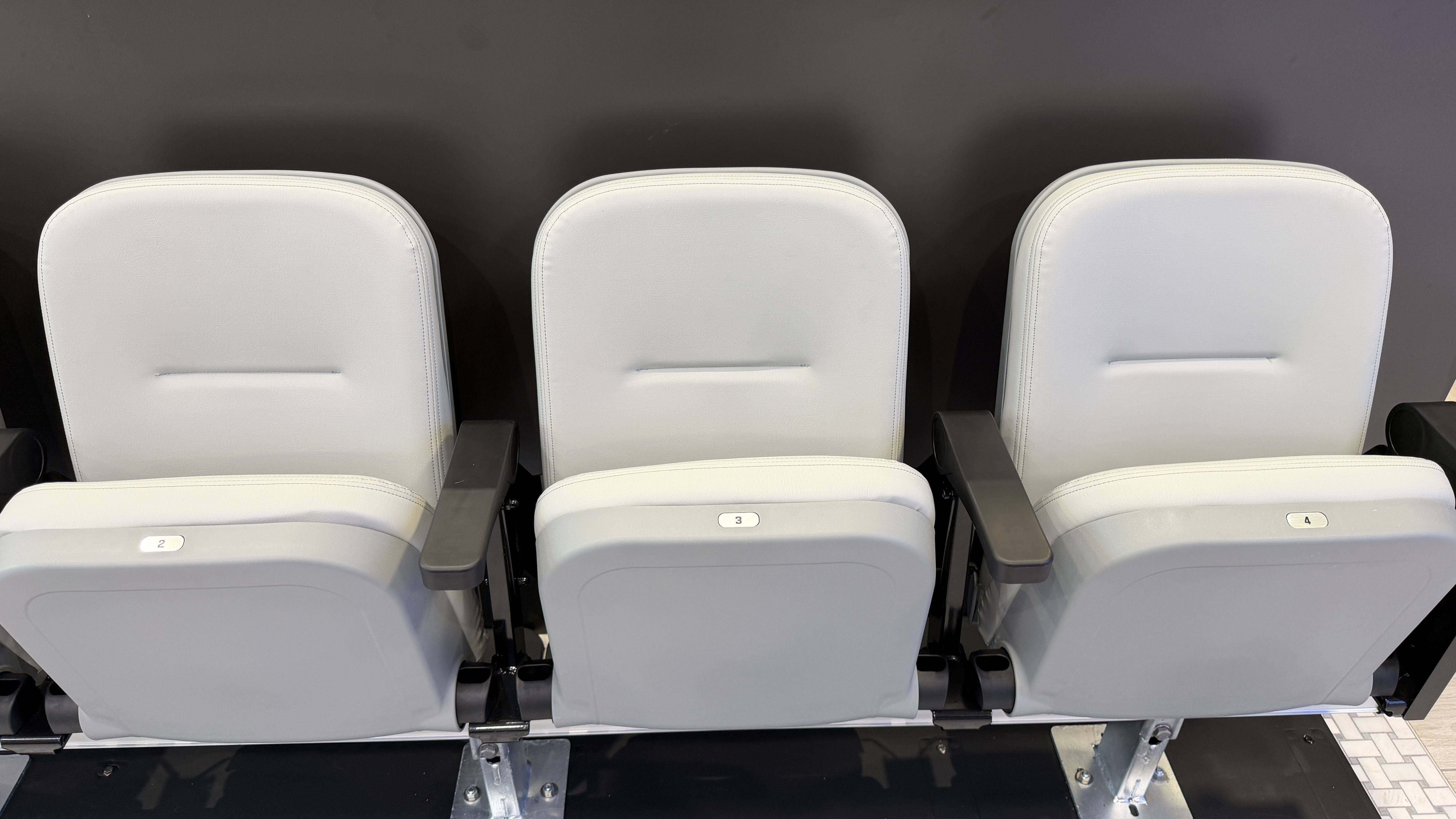 Three light-gray stadium seats with black armrests in a row, against a dark wall; number plaques show 2, 3, and 4.