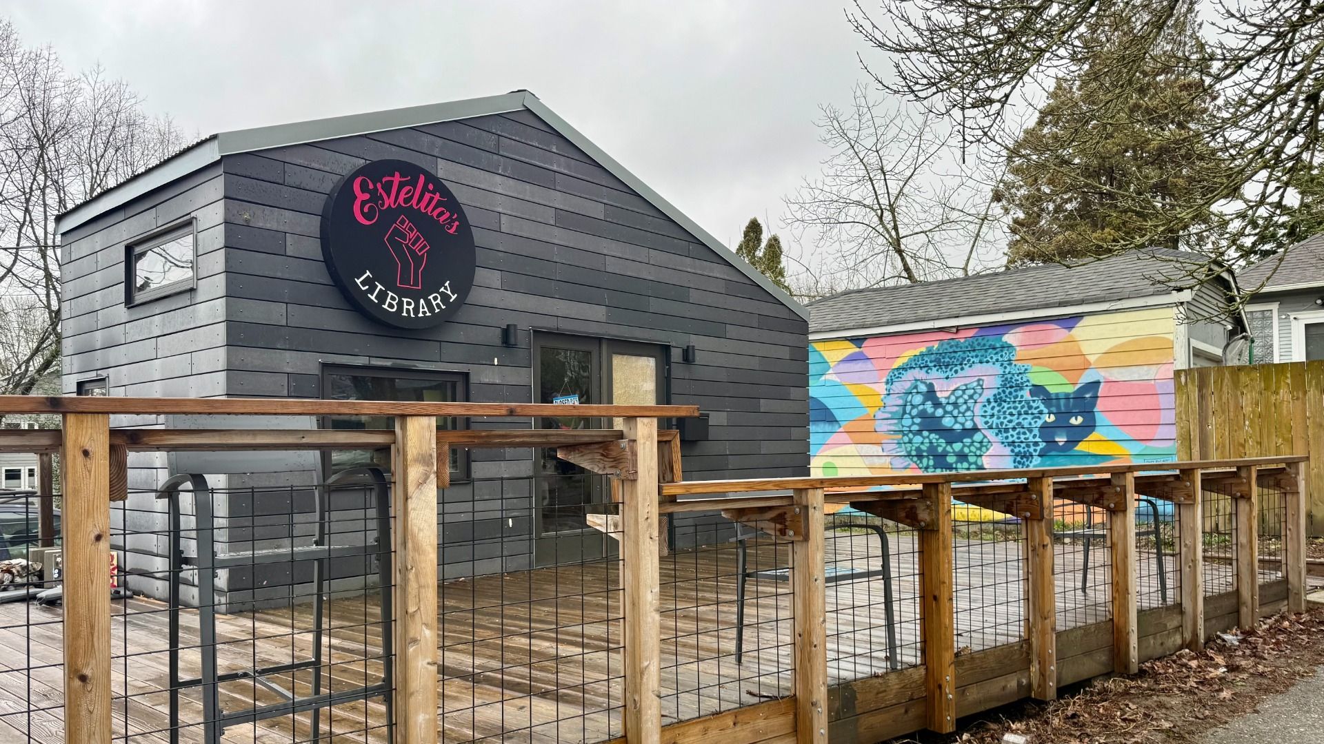 Gray building with black sign reading "Estelita's Library" in pink and white, featuring a raised wooden deck with metal chairs. Colorful mural of blue cat on adjacent wall.