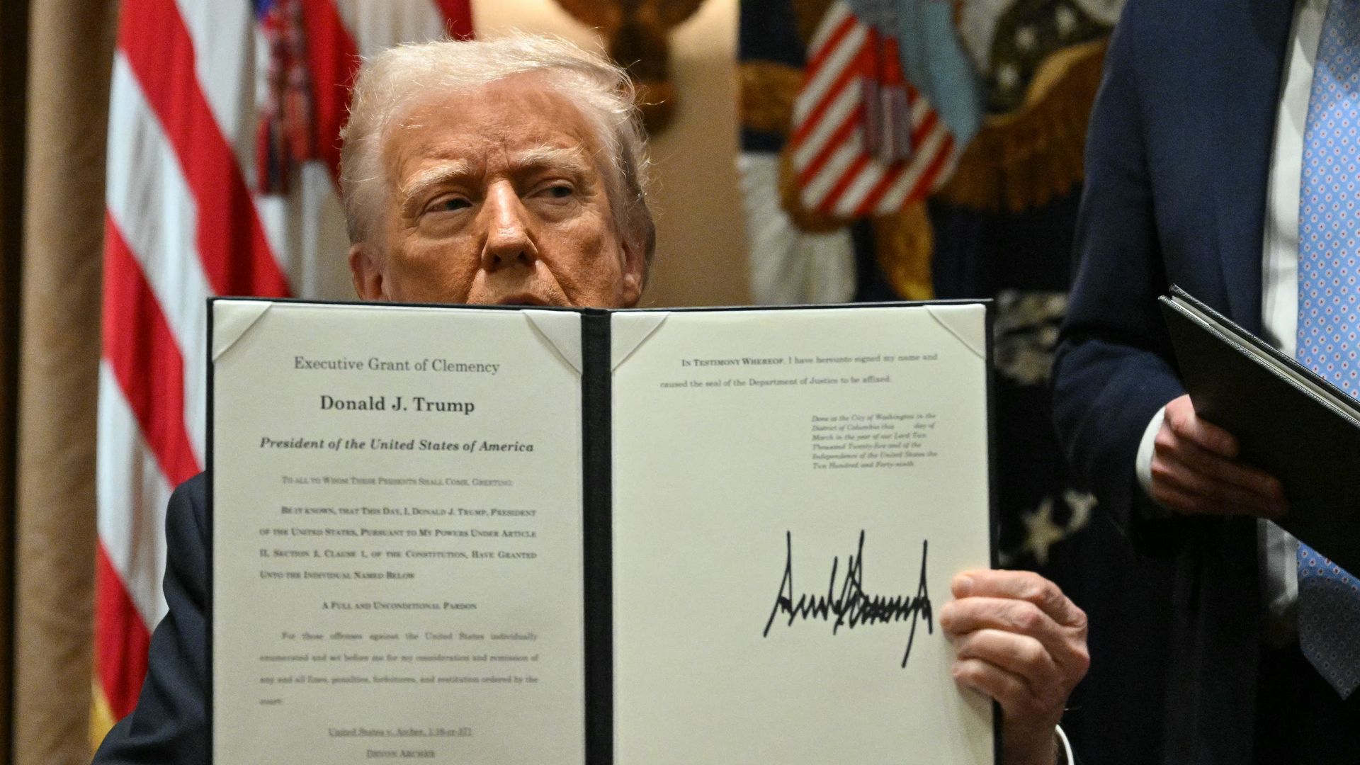 Donald Trump, sitting in front of an American flag, holding an executive grant of clemency that covers part of his face.