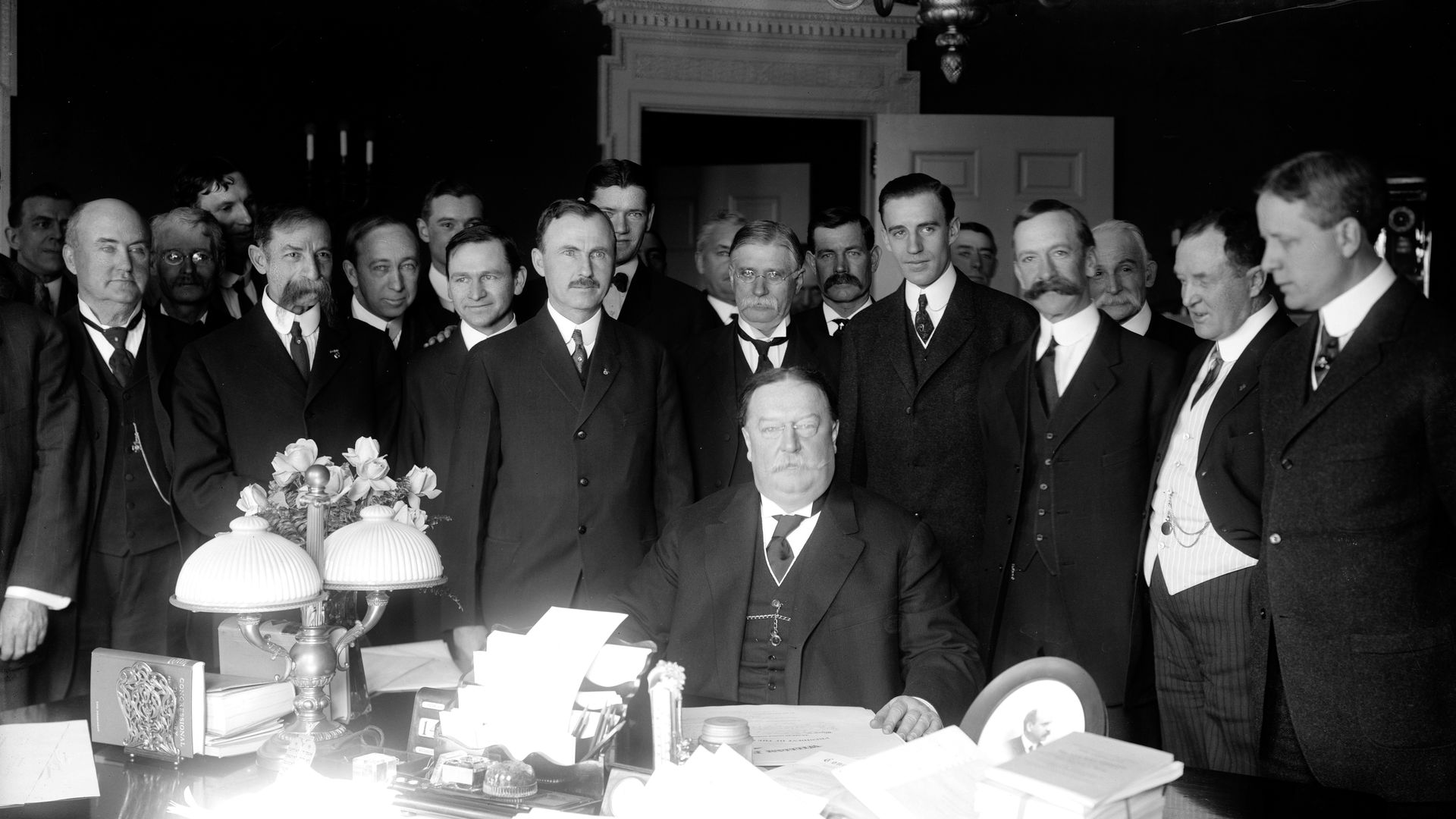 William Howard Taft sits at a desk covered in paperwork while a large group of men stands behind him.