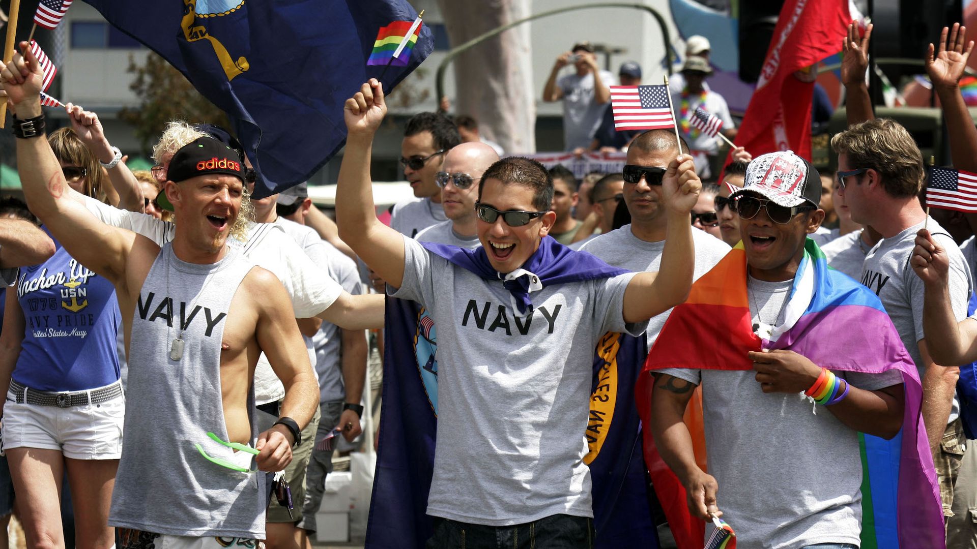 Men wearing NAVY T-shirts march in a pride parade holding flags. 