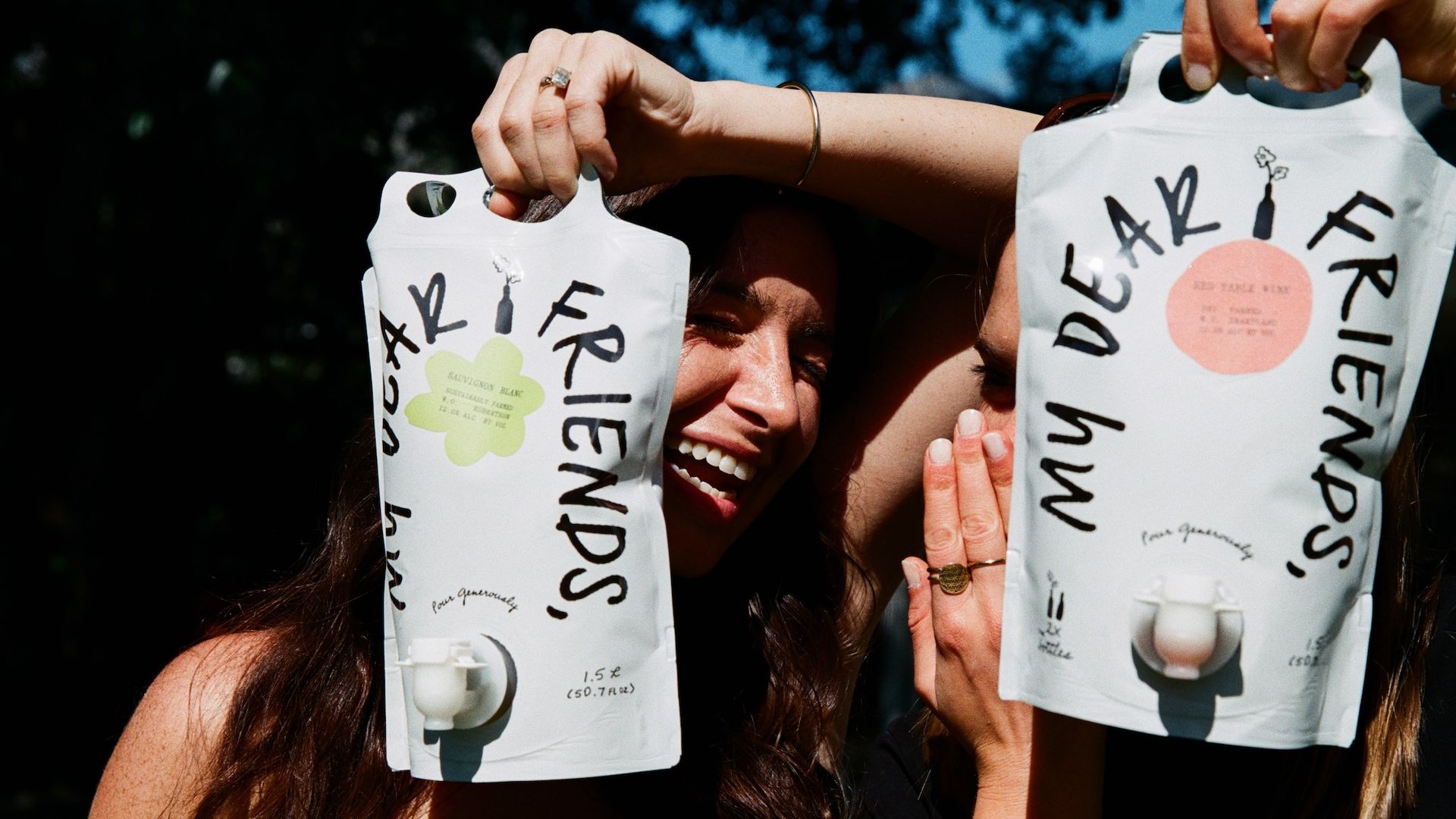 Two people hold up large white bag-shaped containers with bold black lettering and pastel circles, smiling behind them outdoors as sunlight highlights their arms and rings.