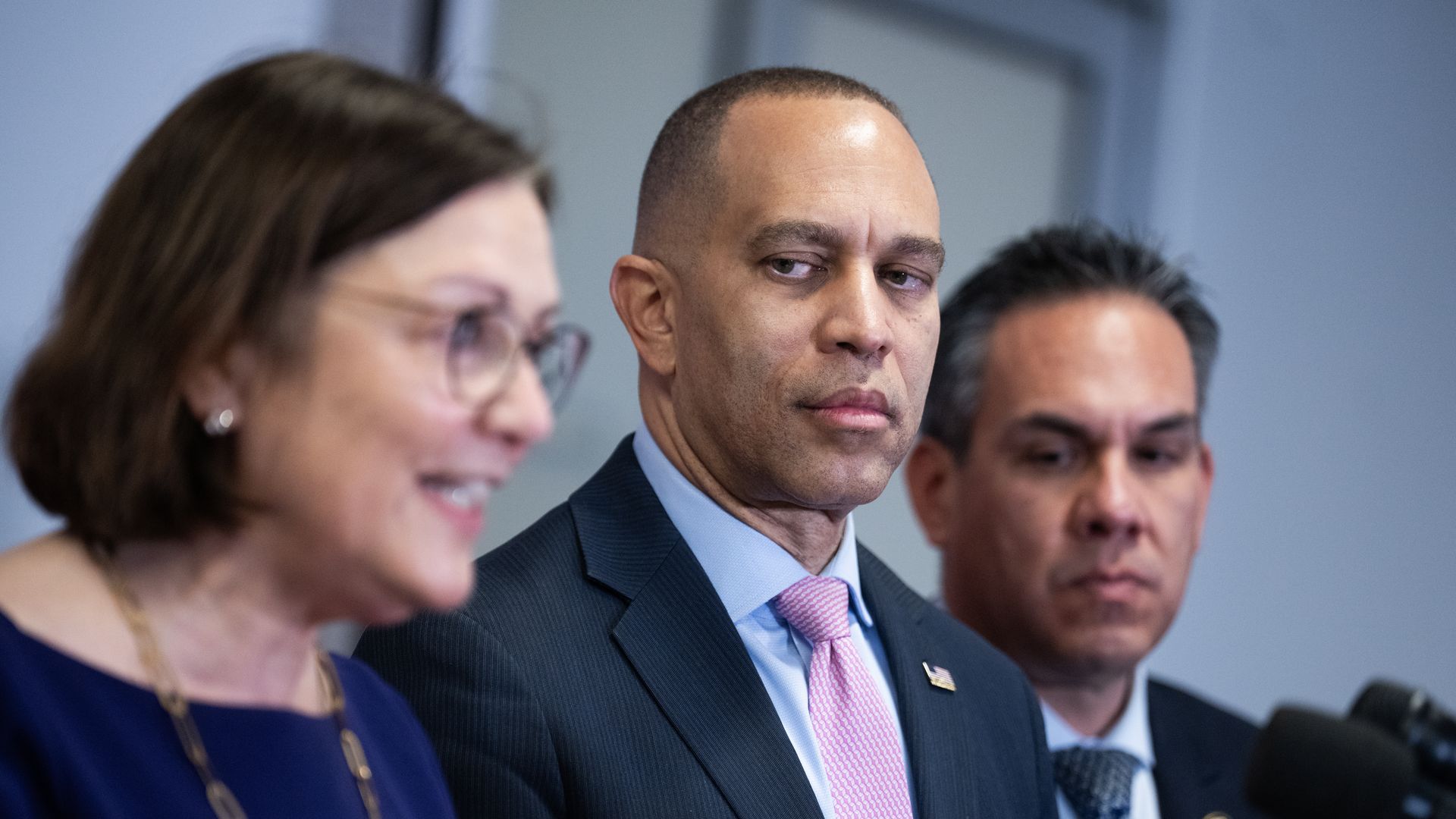 Three people; center is a man in a dark suit and pink tie looking to the right, flanked by a woman in blue on the left and a man in a patterned tie on the right.