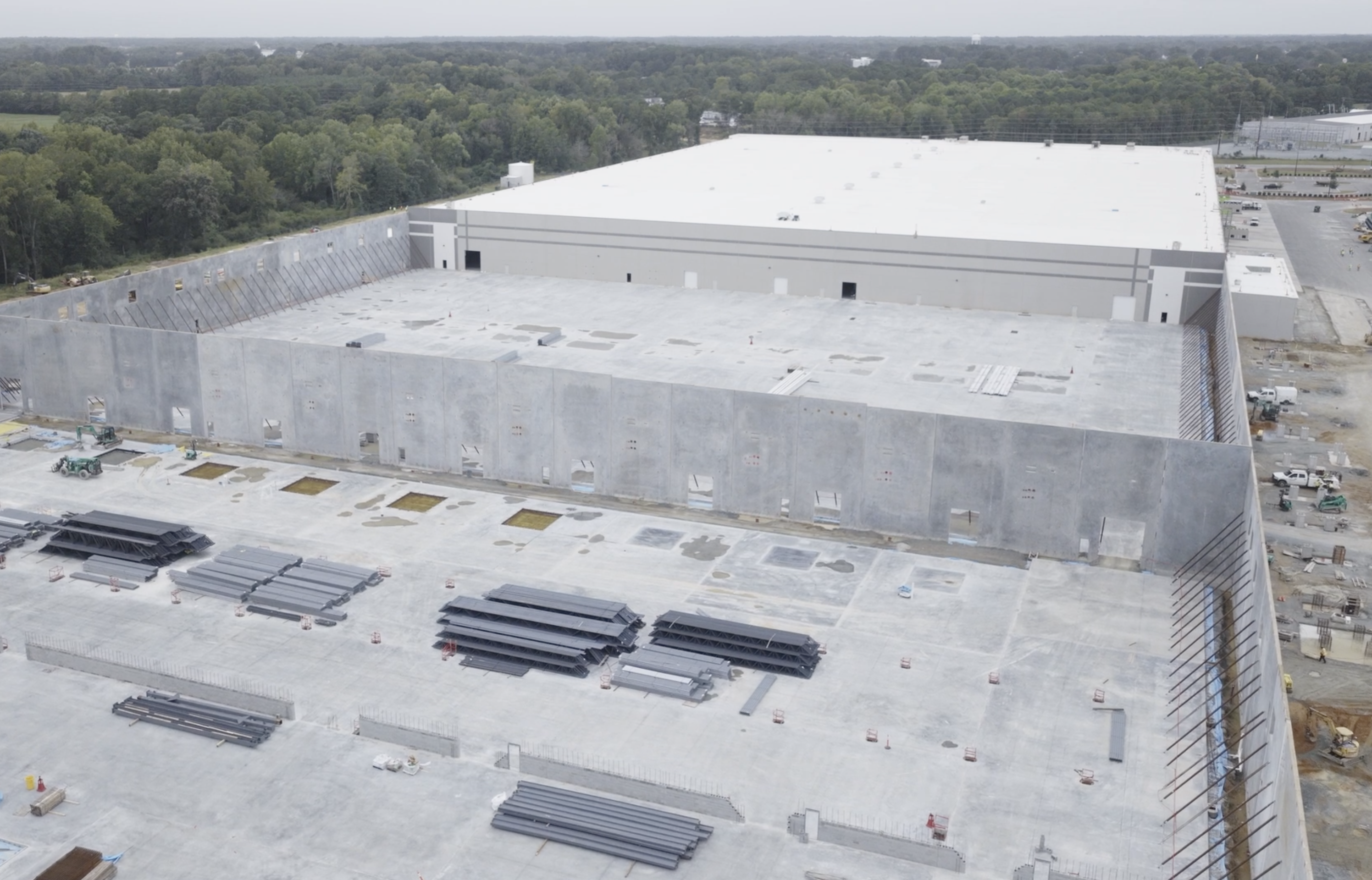 An aerial view shows a large gray warehouse under construction, surrounded by a tall concrete enclosure and a wide concrete yard with stacked steel beams; forested hills in the distance.
