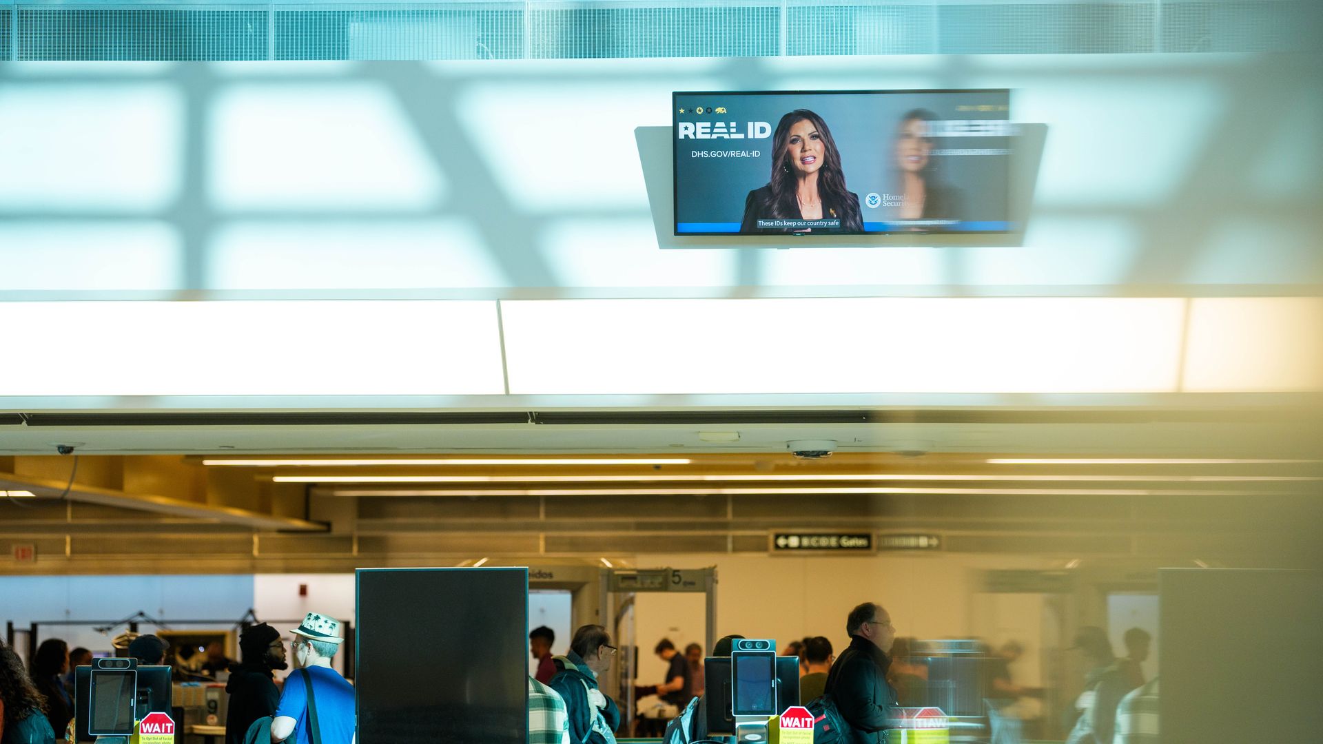 A TV screen plays a video of Kristi Noem speaking about "REAL ID" requirements over the security line at an airport.
