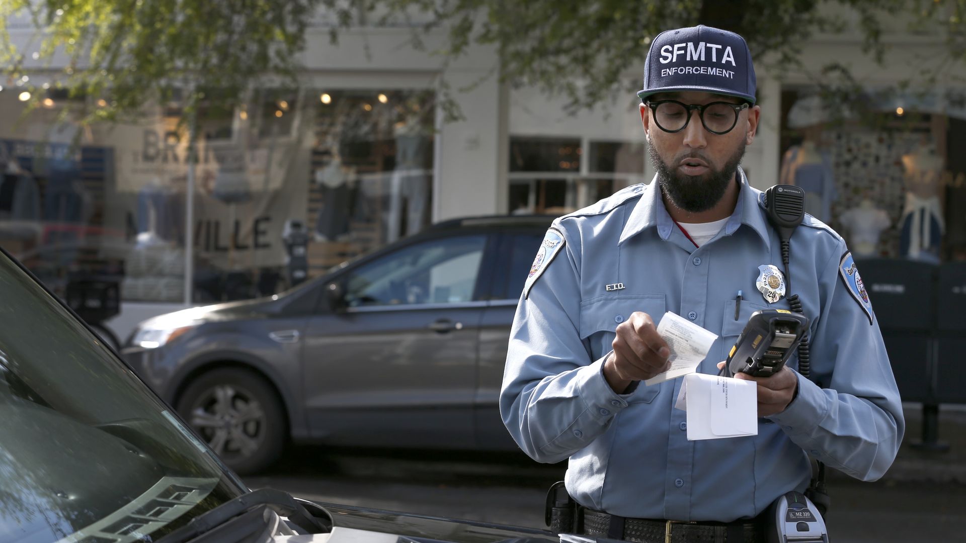 A man in a light blue SFMTA Enforcement uniform is issuing a parking ticket next to a black car on a city street with shops in the background.