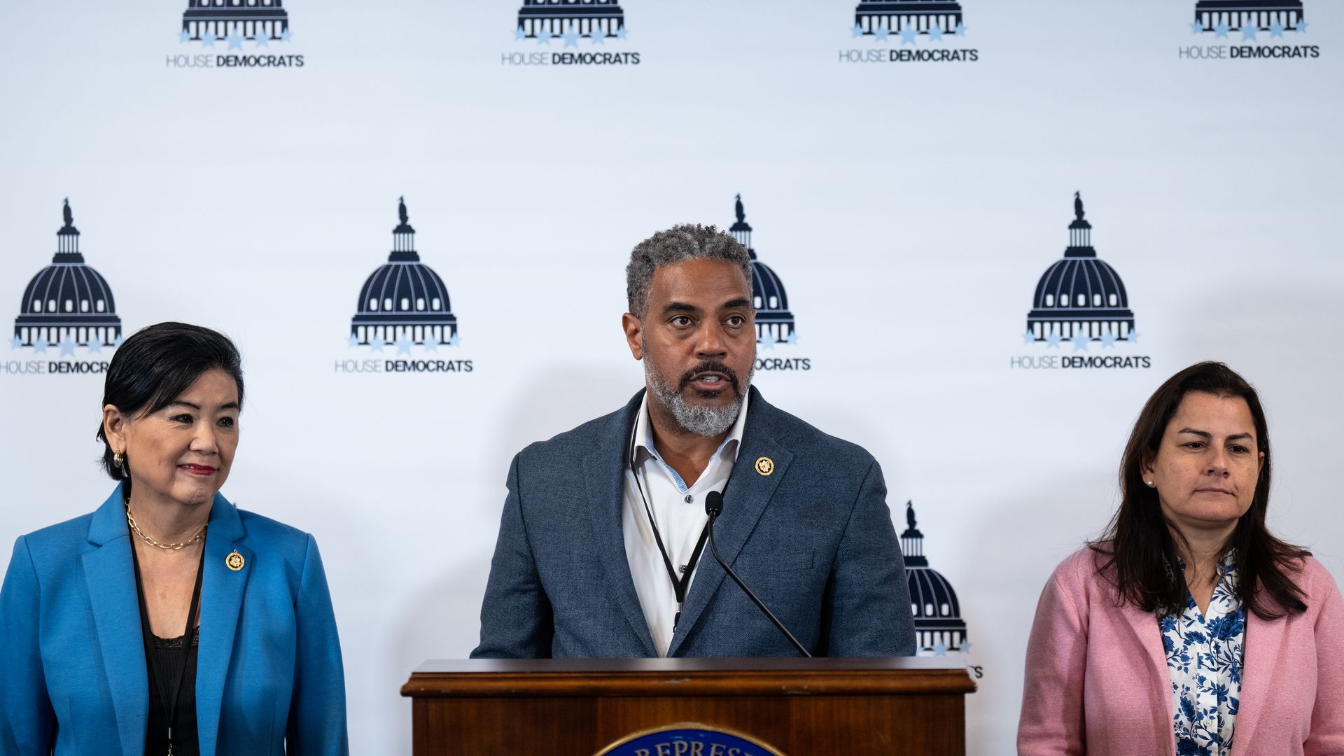 Reps. Judy Chu, wearing a sky blue suit and black shirt, Stephen Horsford, wearing a dark pale blue suit and white shirt, and Nanette Barragán, wearing a light pink suit and white and blue floral shirt, standing in front of a white backdrop with the House Democrats logo.