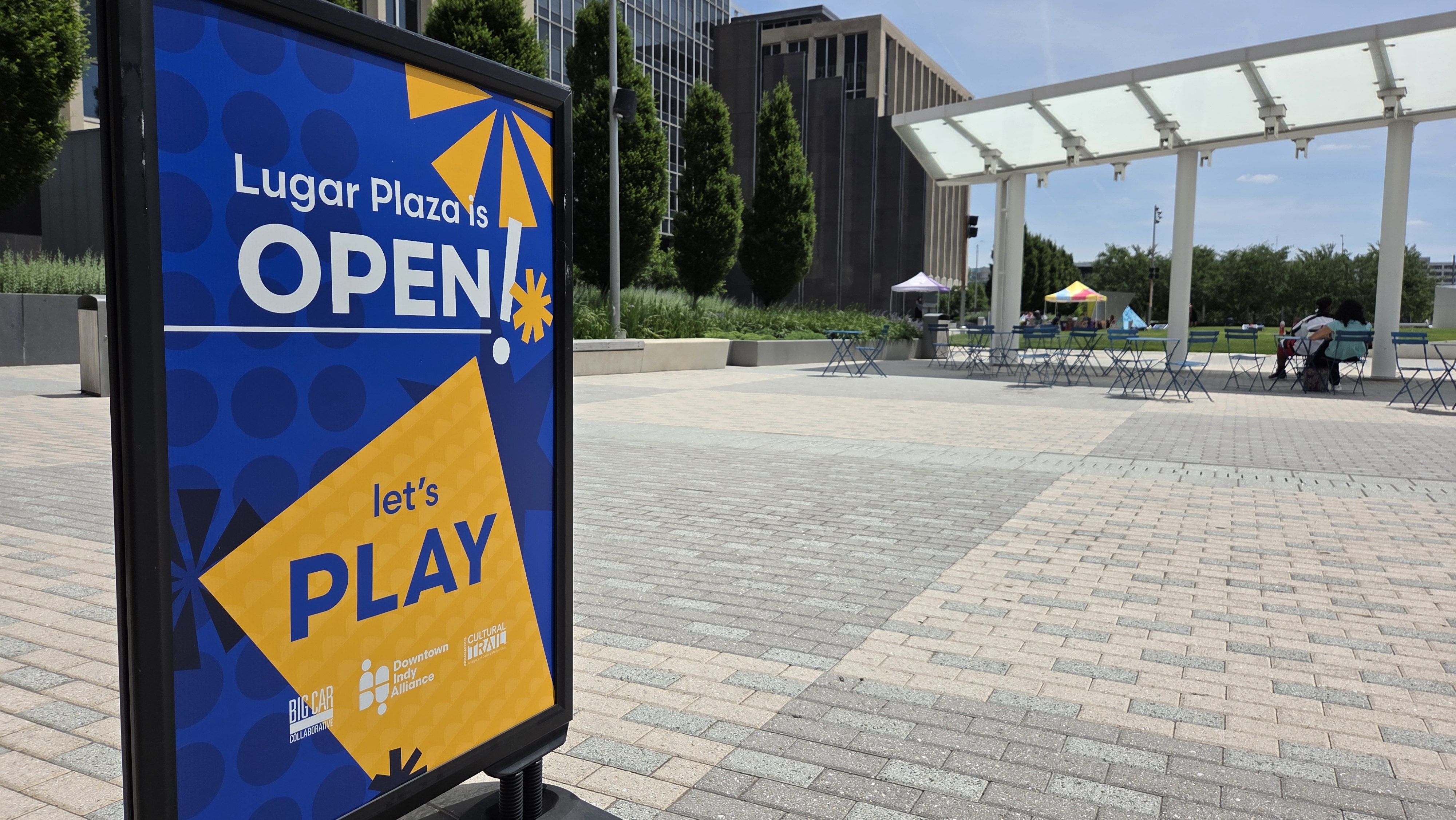 Large blue sign reads "Lugar Plaza is OPEN" with yellow shapes; behind it, an open plaza with a white pergola, green trees, and scattered blue tables and chairs with people sitting.