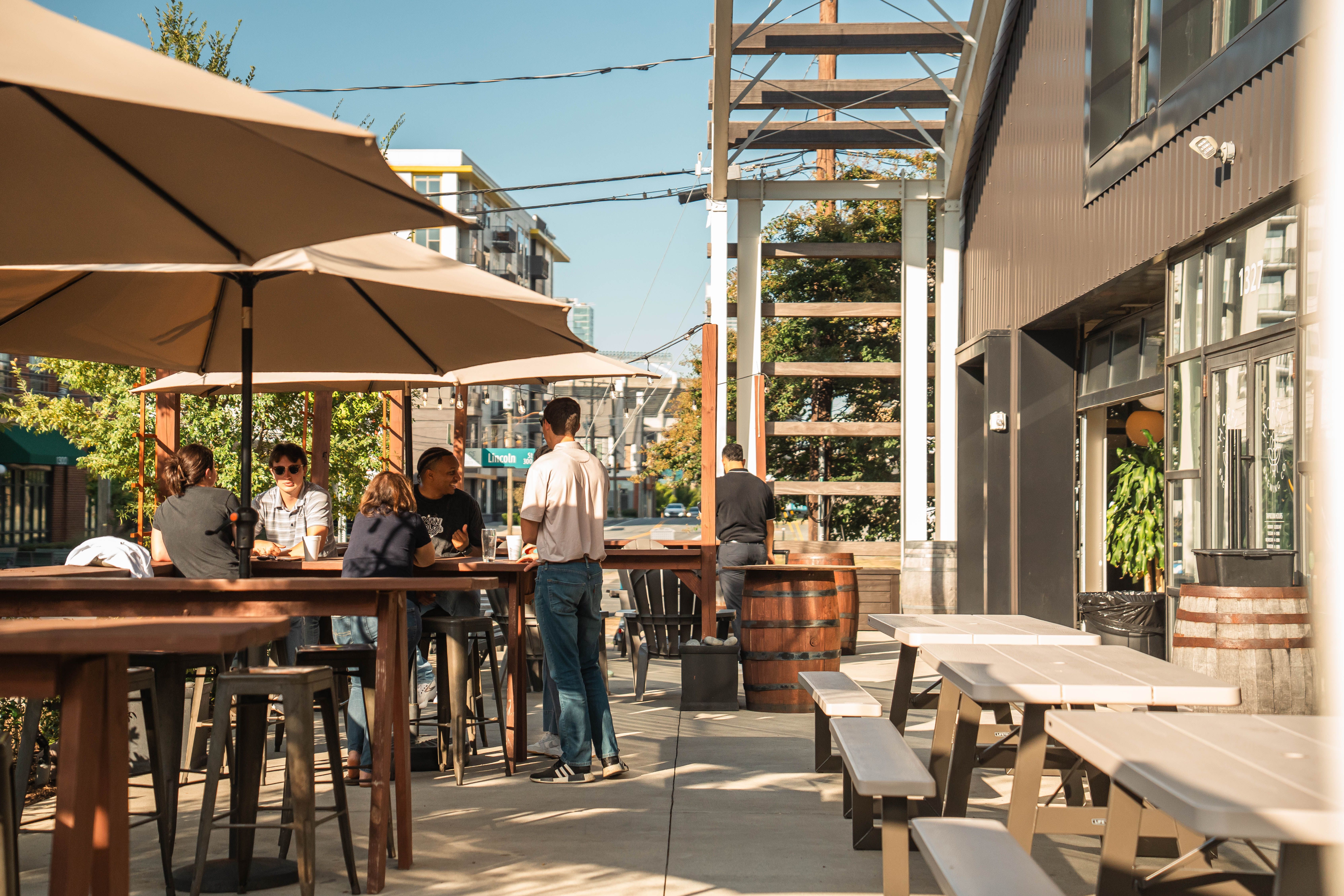 Outdoor patio seating beside a modern building, with guests gathered under large umbrellas at communal tables on a sunny urban street.