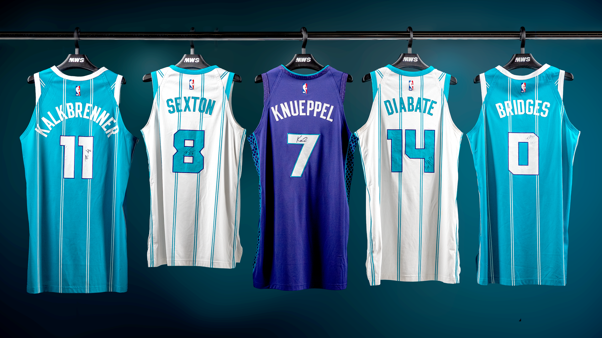 Five basketball jerseys hanging on a rack, three teal with white stripes and two white with teal stripes, names Kalbrewner, Sexton, Knueppel, Diabate, Bridges, all signed.