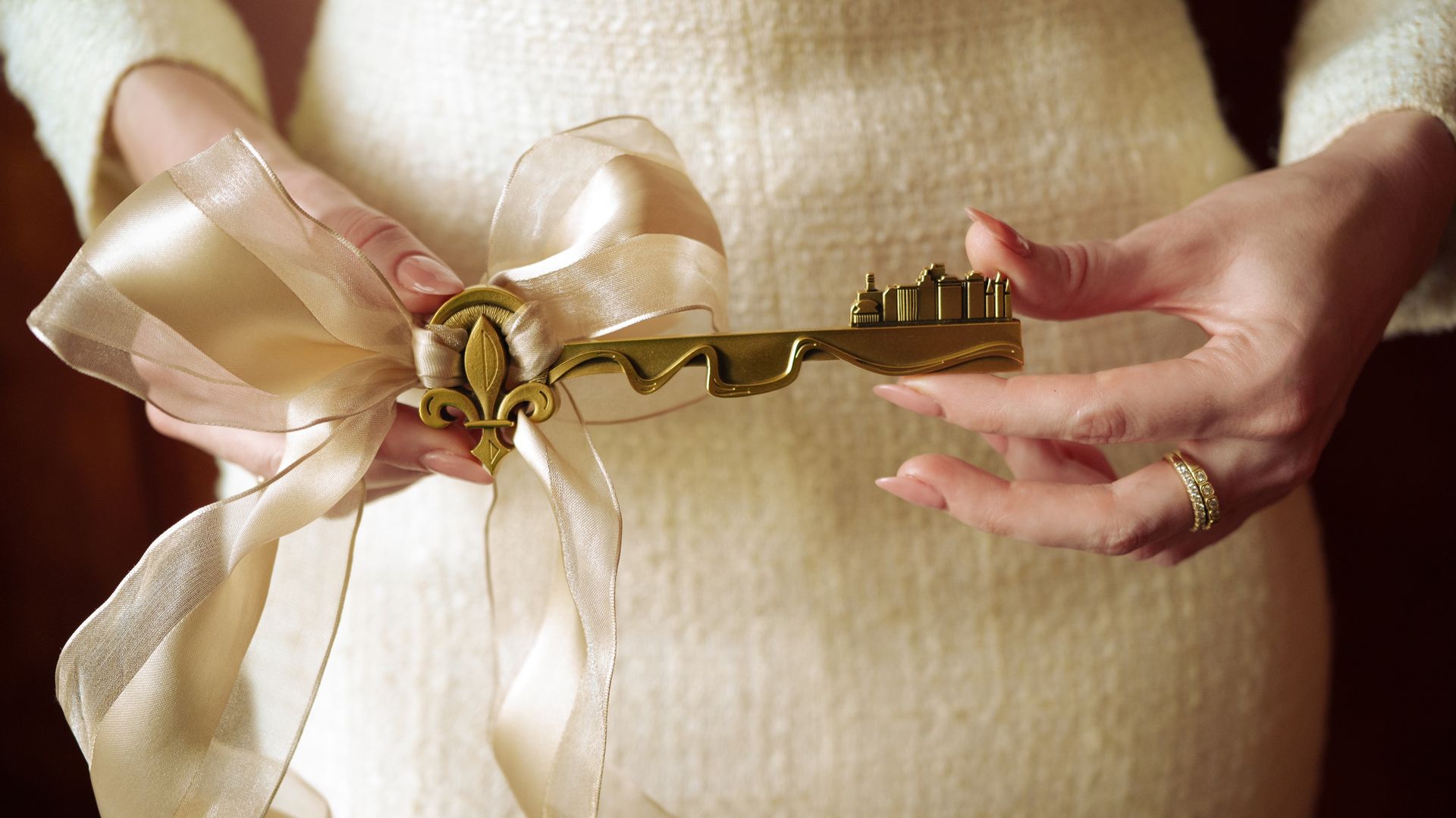 A person in a cream textured sweater holds a large ornate gold key tied with a pale ivory ribbon; the key features a fleur-de-lis motif.