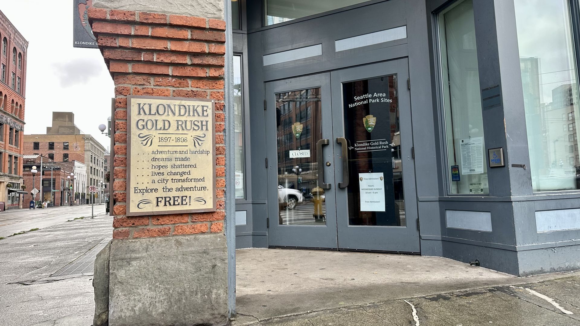 A placard on a brick column says "Klondike Gold Rush" and advertises free exhibits. The National Park Service logo appears on the double doors that open into the building. The doorframes are painted blue; brick buildings are visible across the street.