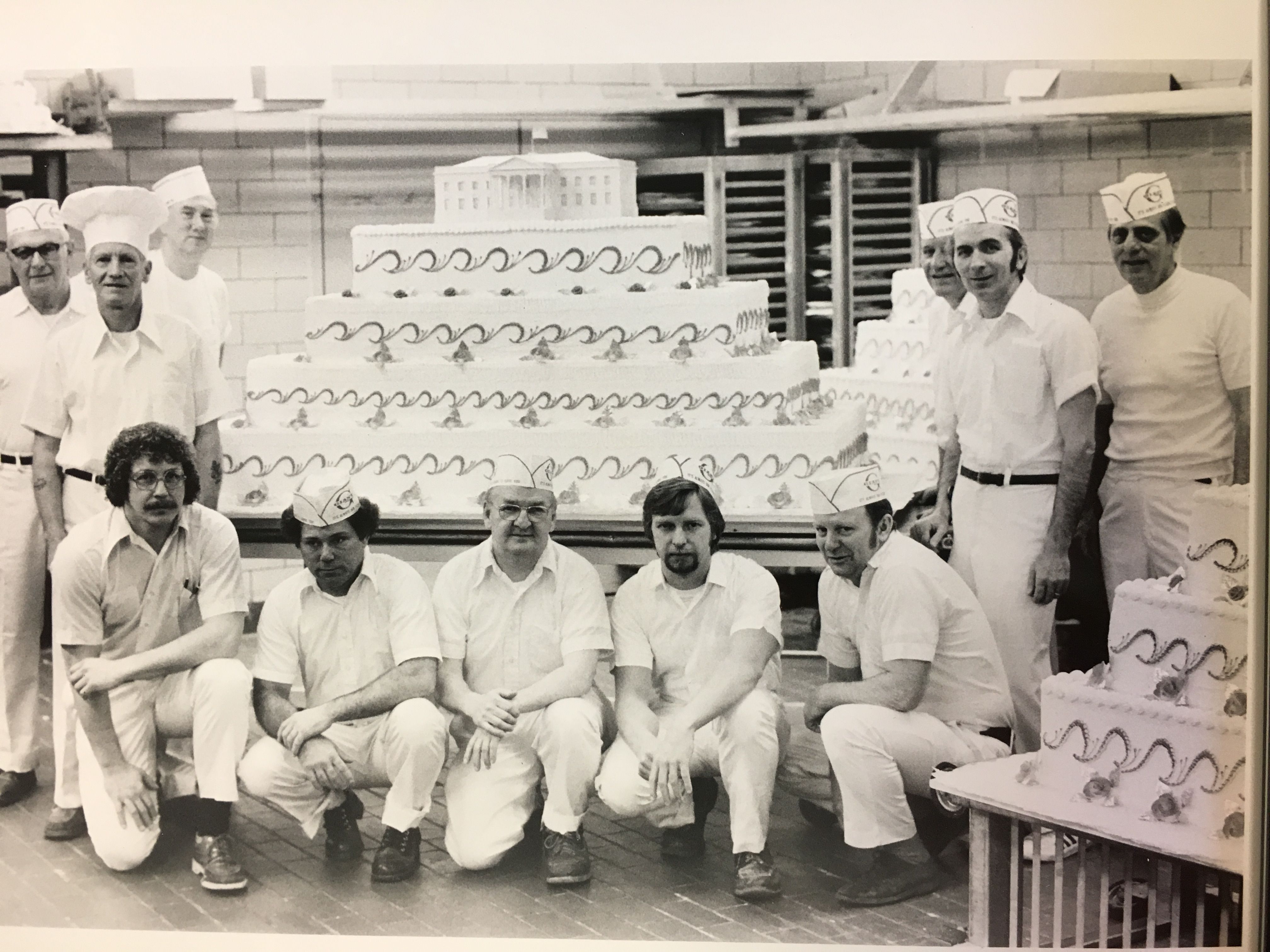 Black and white photo of eleven bakers in white uniforms and hats posing with three large decorative cakes, one topped with a model of a building, in a bakery kitchen.