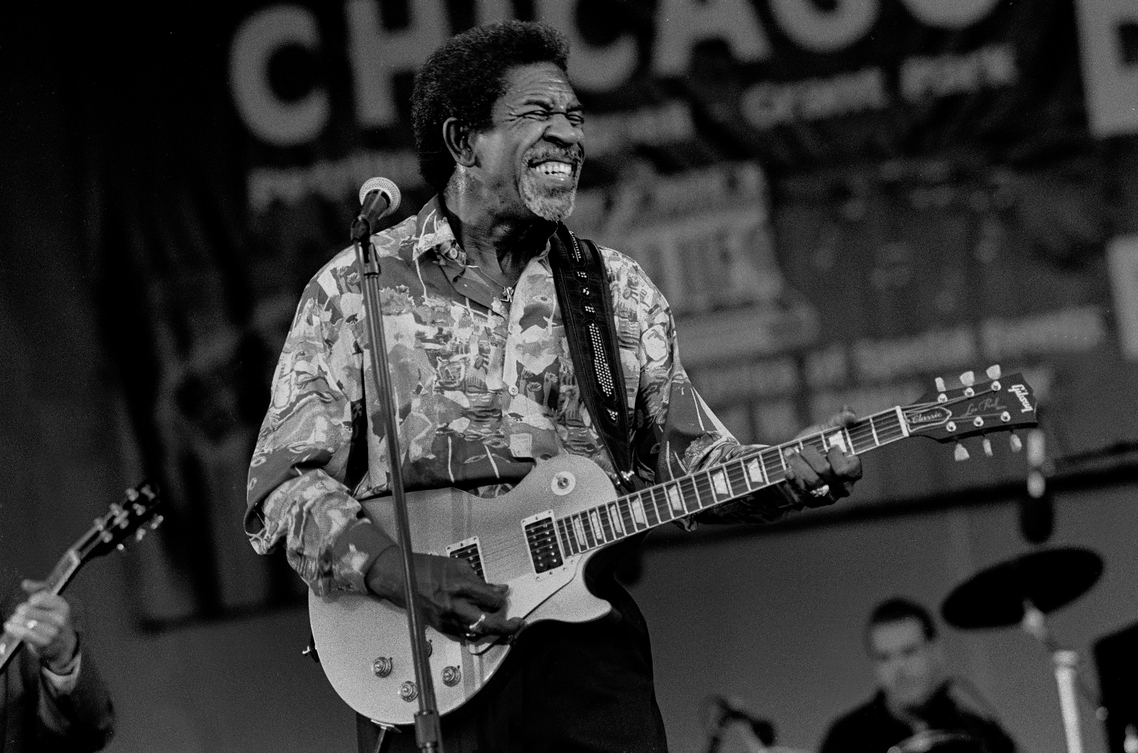Black-and-white photo of an older guitarist on stage, wearing a patterned shirt, smiling while playing an electric guitar near a microphone, with a blurred audience in the background.