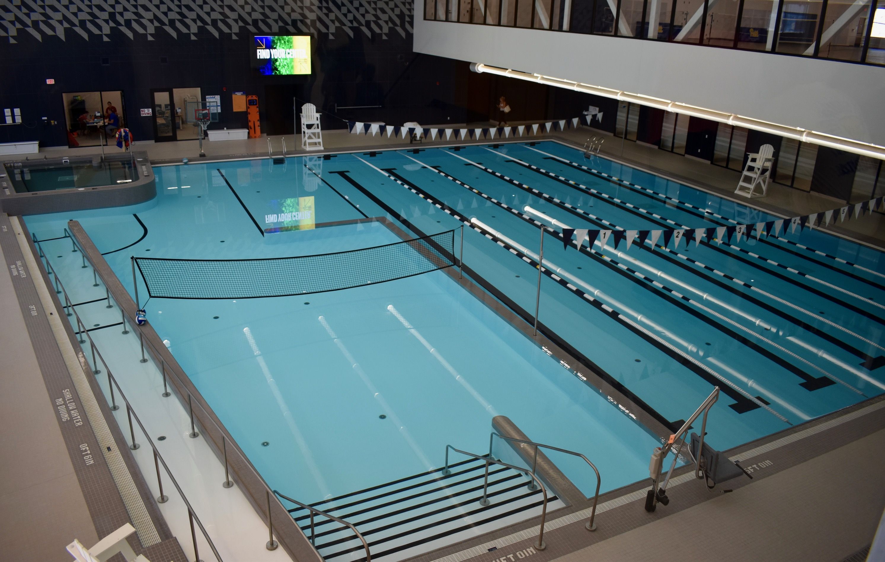 Indoor swimming pool with clear blue water, divided lanes, a volleyball net across the shallow end, lifeguard chairs, and a digital sign reading "FIND YOUR CENTER" on a dark wall.