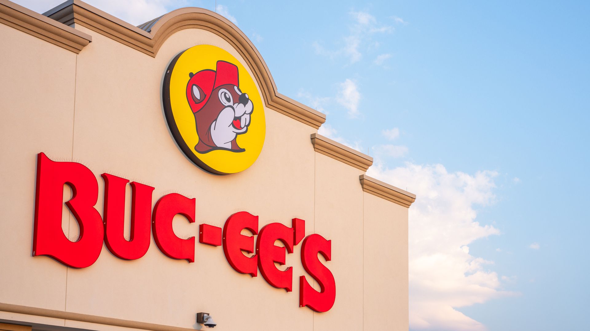Exterior of Buc-ee's store: beige wall with large red letters and a round yellow logo featuring a cartoon beaver in a red hat, set against a blue sky.