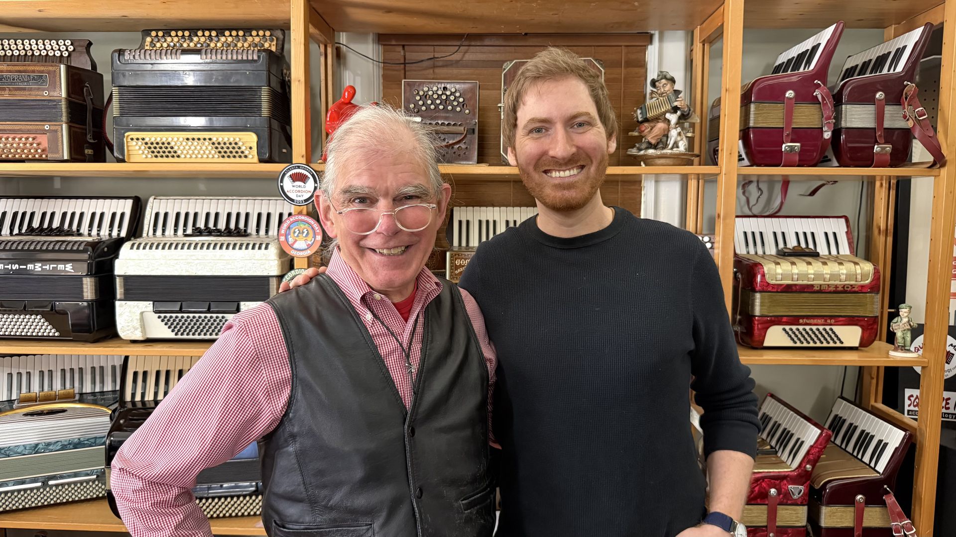 Two men smile in a shop filled with accordions on wooden shelves; one wearing a red check shirt and black vest, the other in a black sweater, with many vintage instruments behind them.