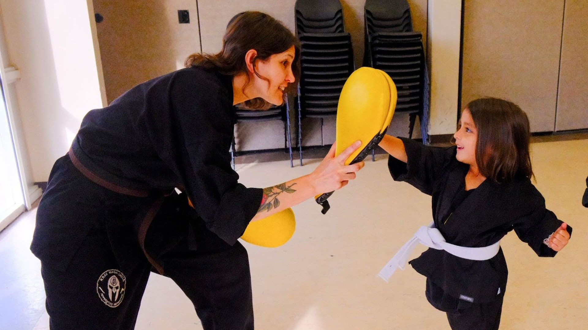 A photo of an adult martial arts instructor holding up a yellow punching bag for a child martial artist to hit
