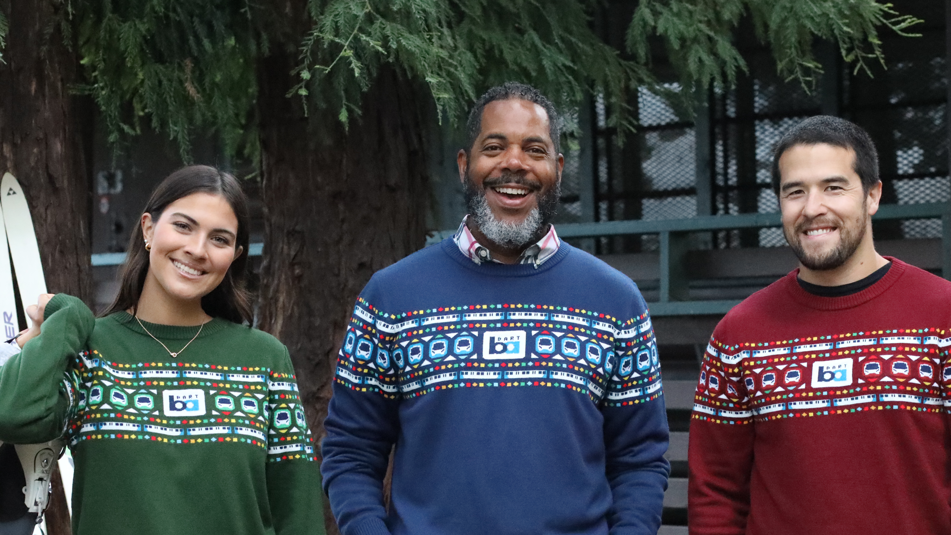 Three smiling people wearing festive sweaters with colorful BART train patterns: a woman in green, a man in blue, and a man in red, standing outdoors in front of trees.