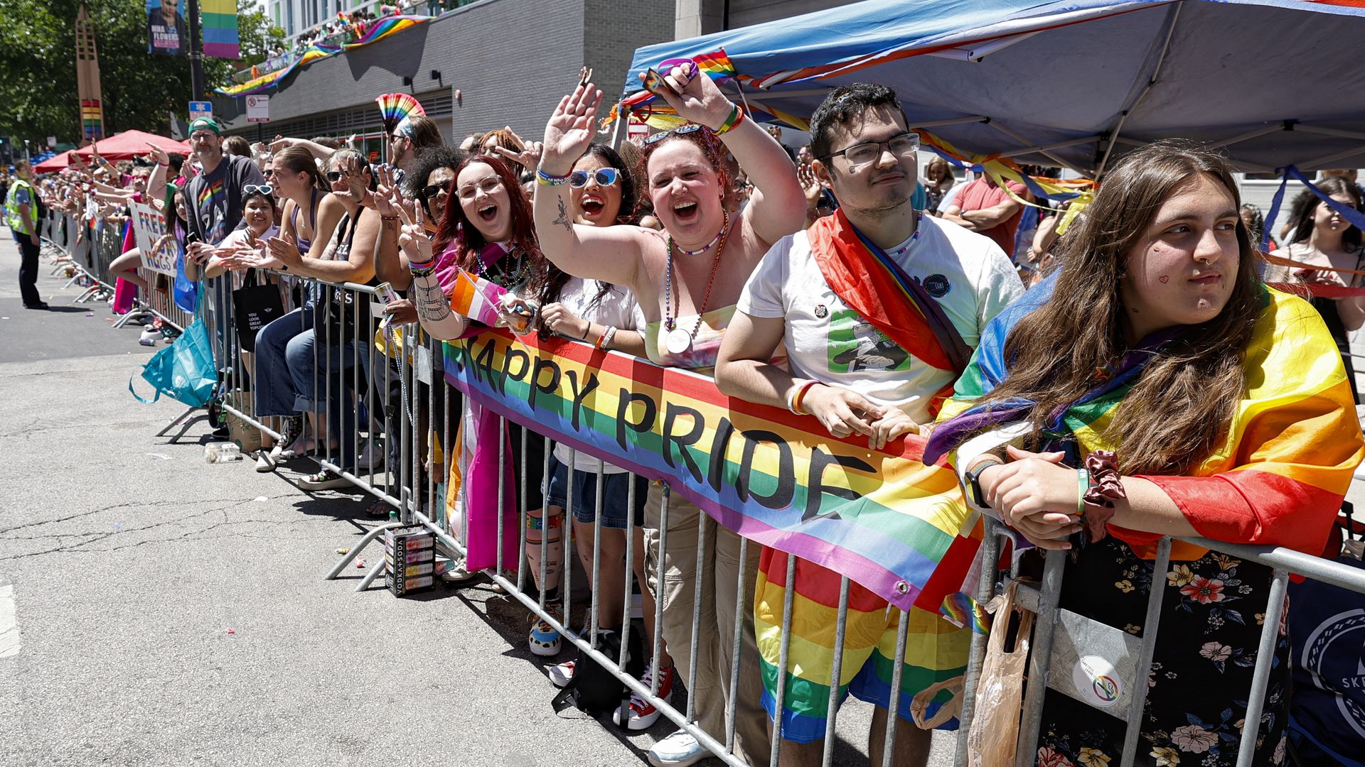 Cheering crowd along a city street at a Pride parade, waving rainbow flags and banners in colorful outfits, under a blue canopy over barricades.