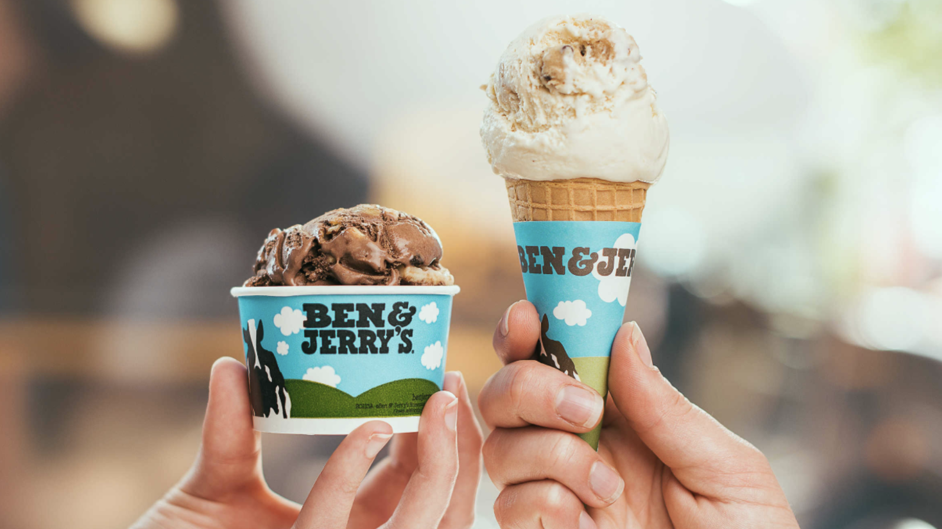 Two hands hold Ben & Jerry's: a cone with vanilla ice cream and a cup of chocolate ice cream, blue wrappers with clouds and mountains.