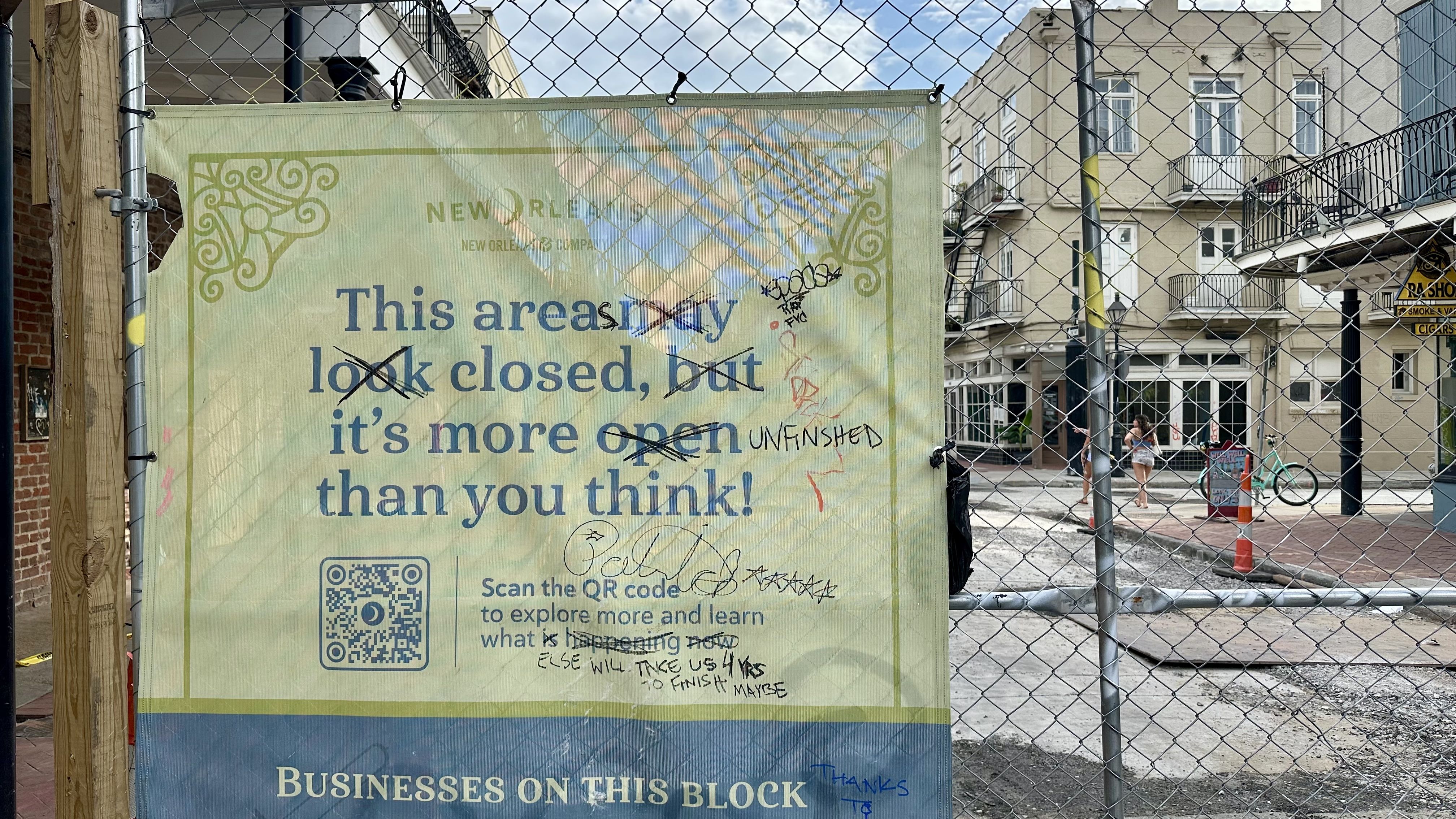Construction banner behind a chain-link fence. It reads, 'This area may look closed, but it's open' with a green decorative border. In the background are beige buildings, a person, and a bicycle.