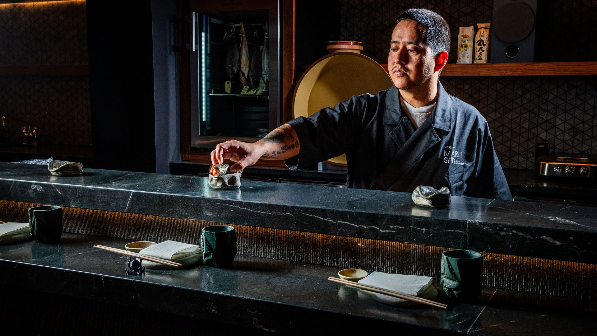 A chef in a dark jacket with "MARU SAI" embroidered serves sushi at a dimly lit stone counter with chopsticks, green cups, and napkins in a modern restaurant setting.
