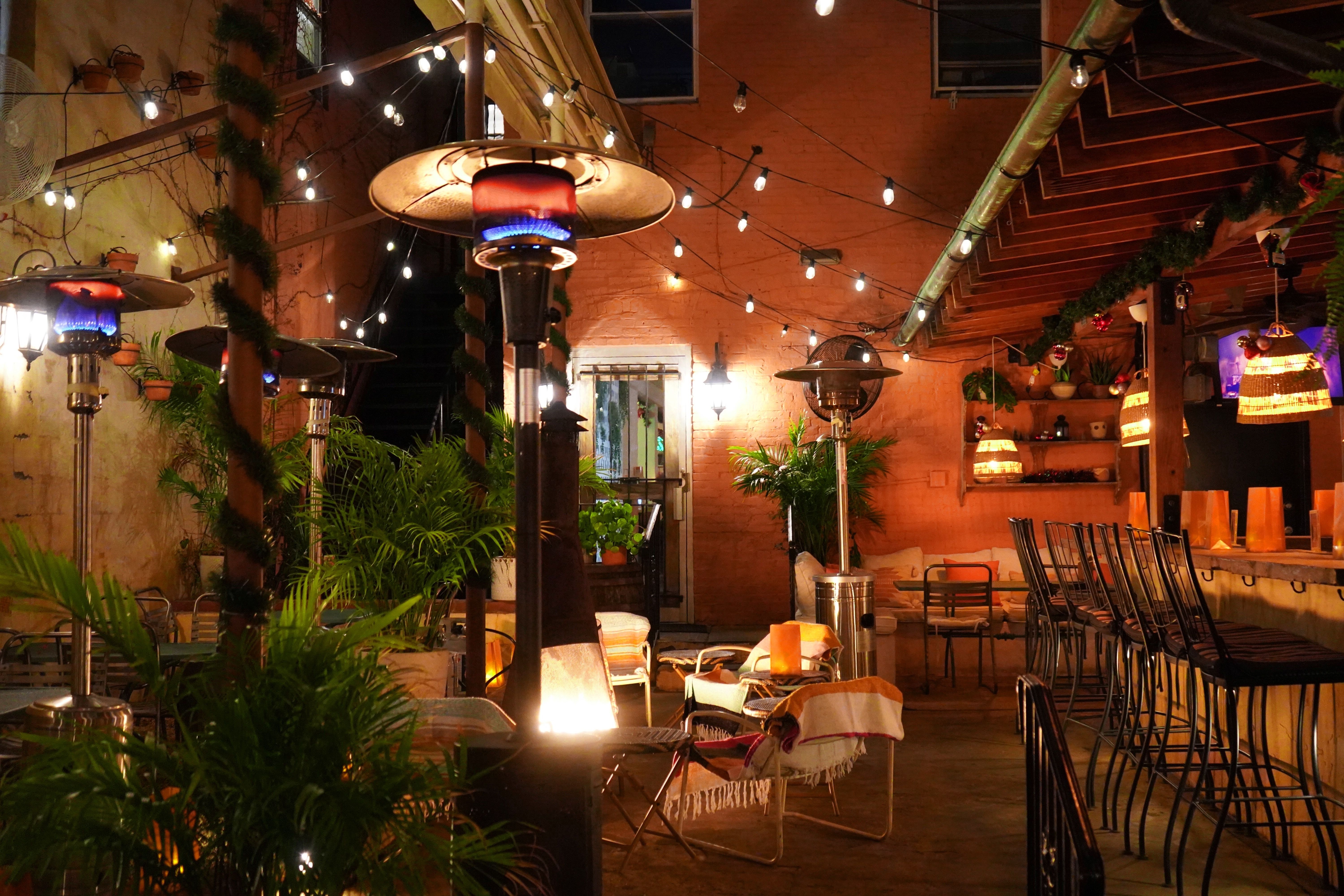 Cozy outdoor patio at night with string lights, tall heaters with blue flames, green plants, wicker chairs with blankets, and a bar area with high stools under warm hanging lamps.
