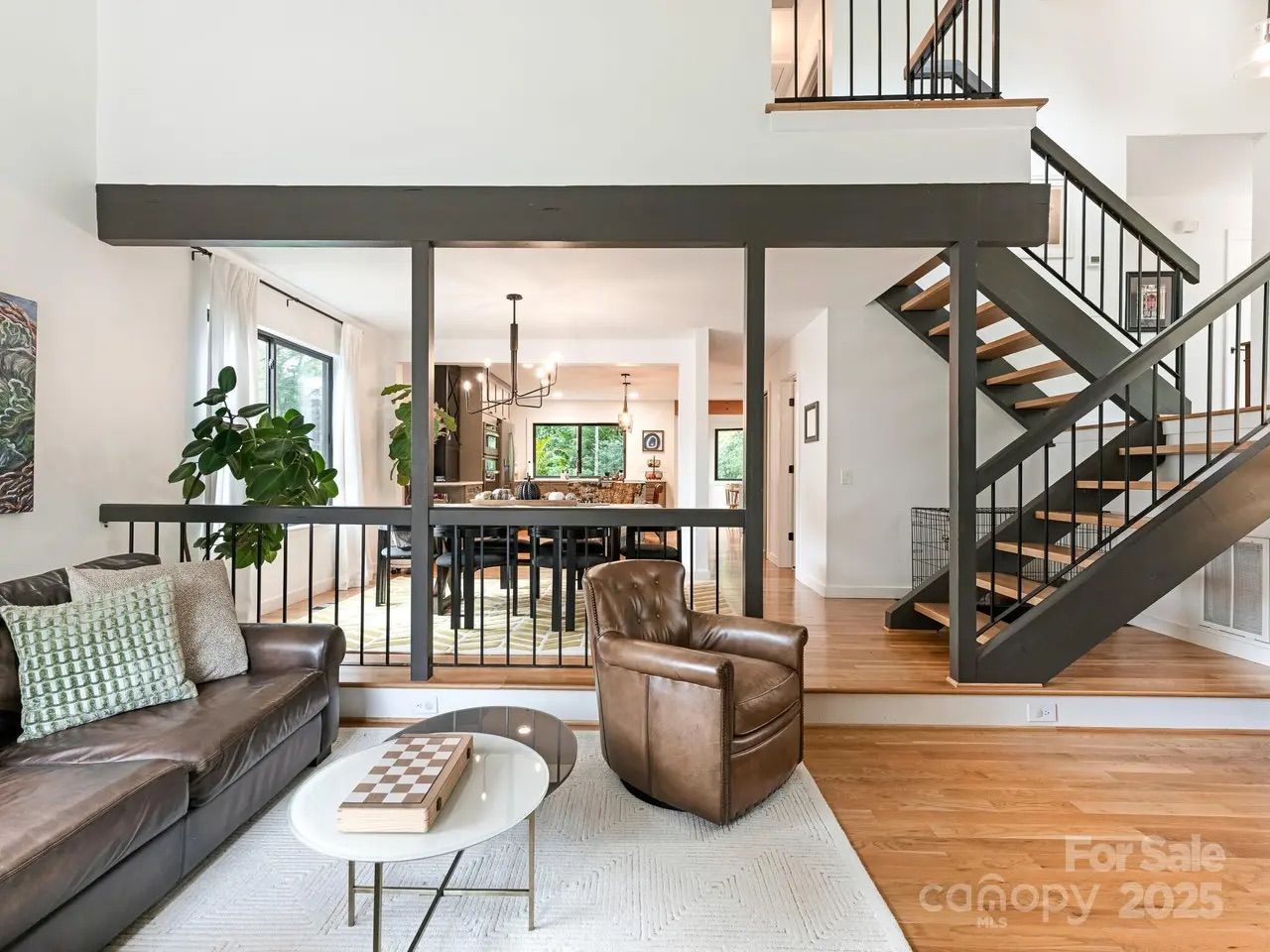 Modern living room with brown leather sofa and chair, white rug, glass coffee table with chessboard, black stairs with wood steps, black railings, and large windows letting in natural light.