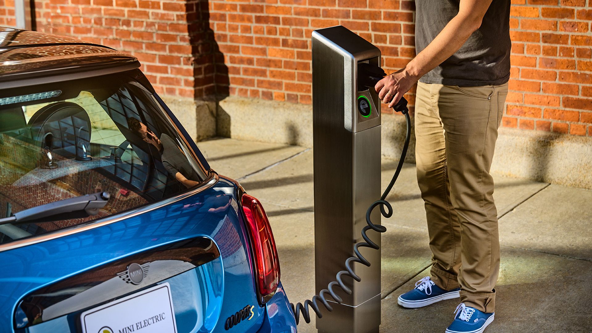 Person in a gray shirt, khaki pants and blue sneakers plugs a cable from his blue MINI electric car into a curbside EV charging pedestal beside a brick wall. 
