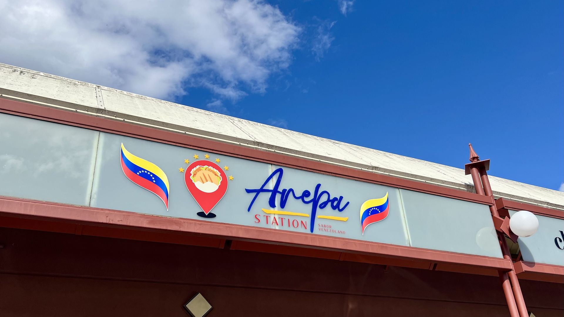 Sign on a building under blue sky reads "Arepa Station" with colorful Venezuelan flags and an arepa icon, indicating a Venezuelan food place.