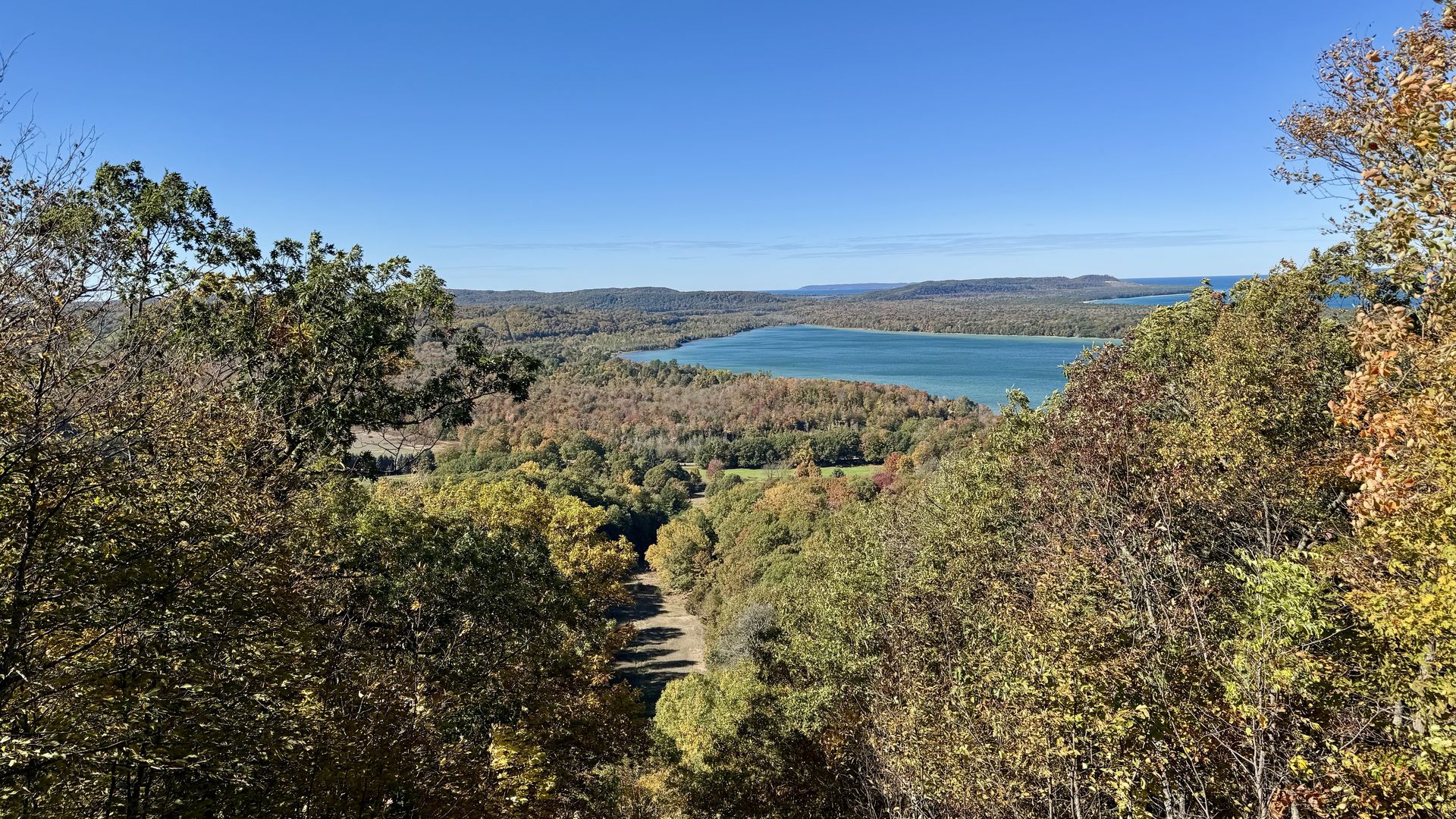 Panoramic view over a wooded valley to a blue lake; autumn foliage in foreground, a dirt path winding down, and distant hills under a clear blue sky.