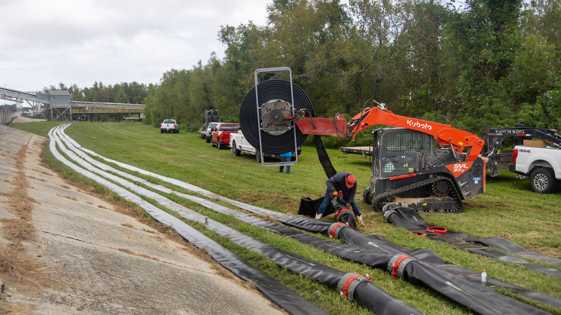 Rows of flexible pipeline are laid on the batture. Construction equipment waits nearby.