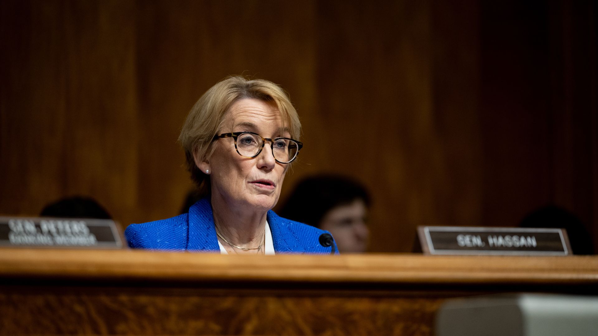 A woman with short blonde hair and glasses wearing a bright blue blazer speaking at a wooden desk with a microphone, dark blurred background with nameplates showing Sen. Hassan.