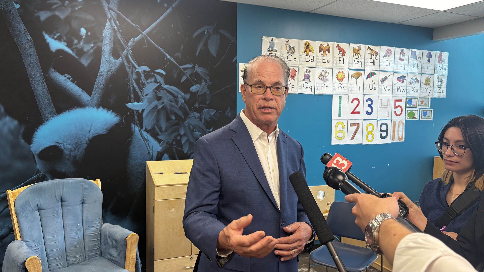 A man in a blue suit speaks to reporters in a room with a bright blue wall; reporters hold microphones, and the wall is decorated with alphabet charts and large colored numbers, plus a blue forest mural on the left.