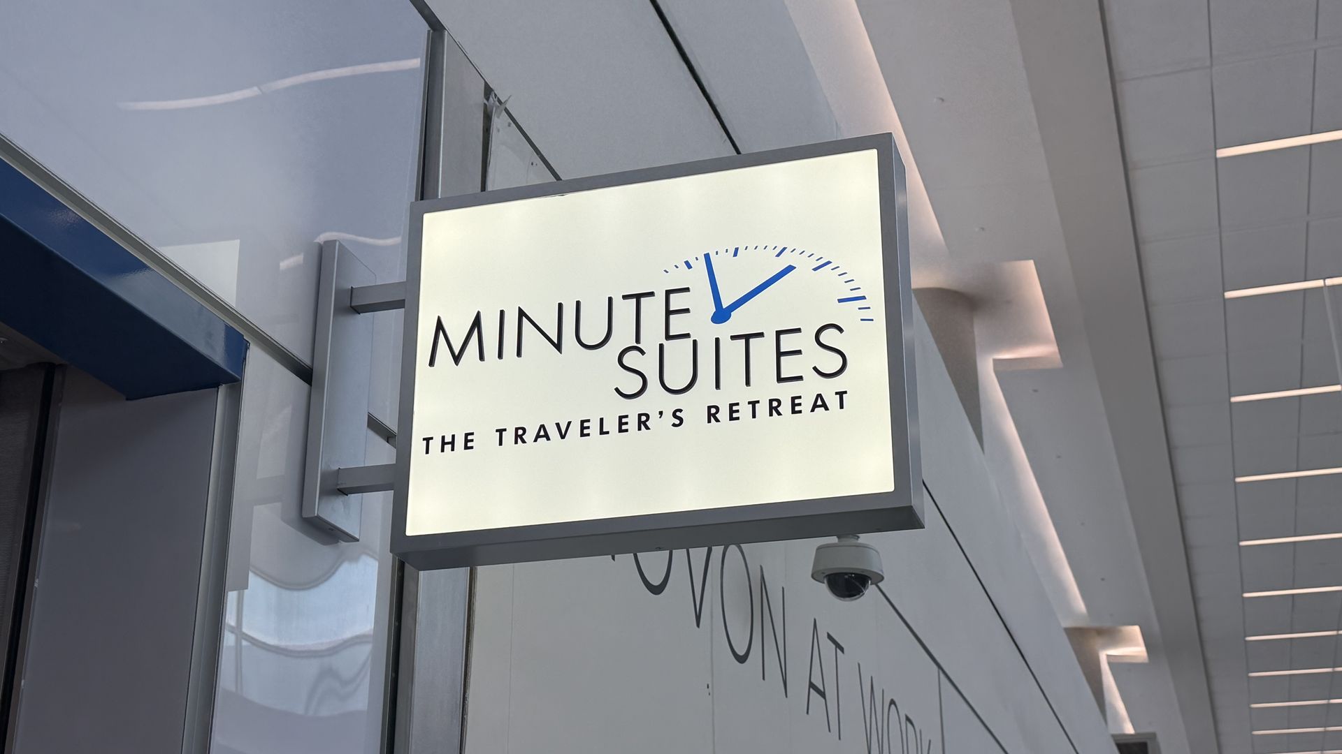 White rectangular sign reading "MINUTE SUITES" with a blue clock graphic; subtitle "THE TRAVELER'S RETREAT". Mounted on a gray metal frame beside glass walls in a modern interior.