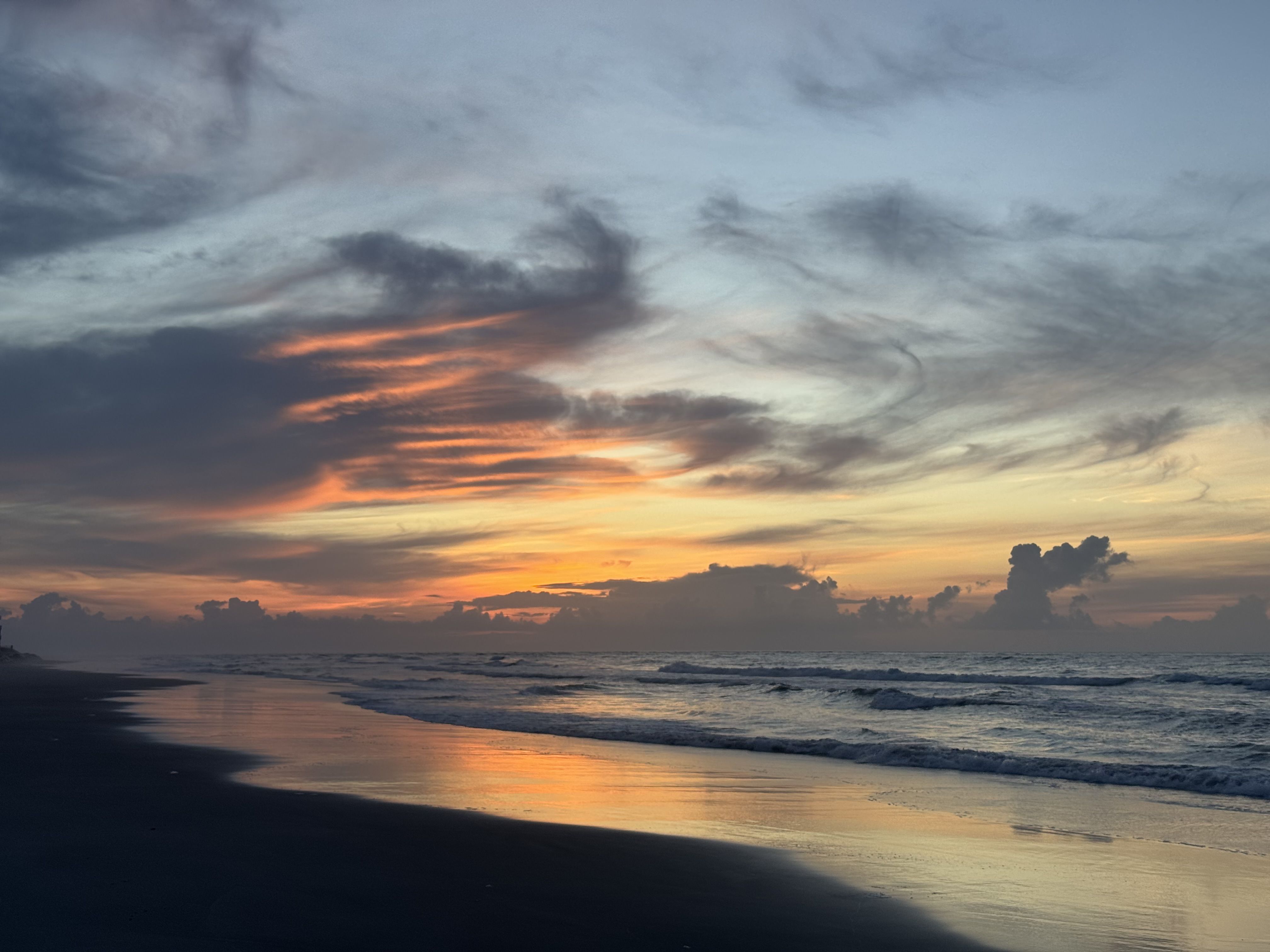 Sunset over a calm ocean with a sandy beach foreground, reflecting orange and pink clouds on wet sand; blue sky darkens to dusk with scattered dark clouds near the horizon.
