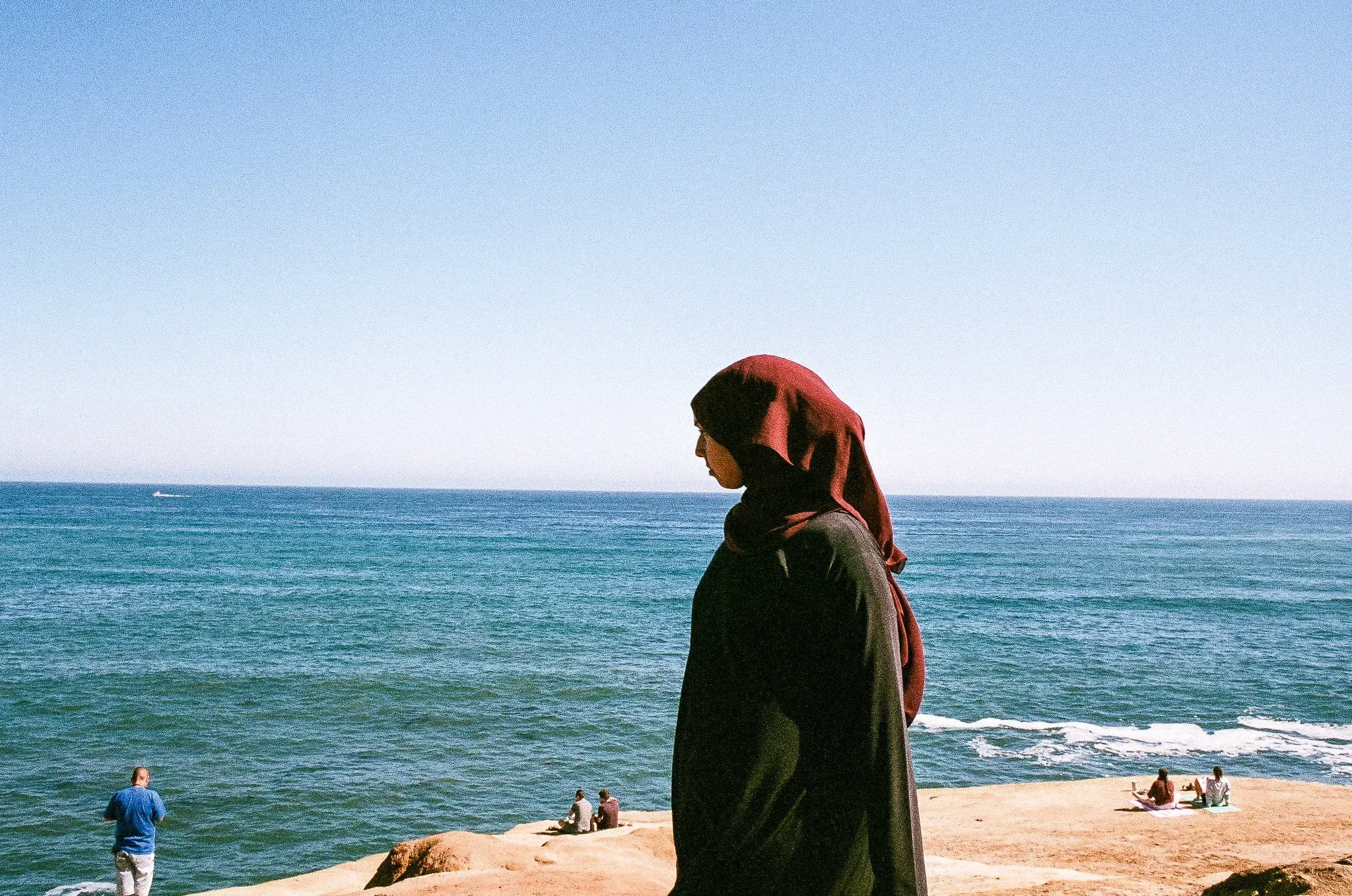 Profile of a woman wearing a red hijab and black clothing standing on a sandy cliff overlooking a blue ocean under a clear sky, with people sitting and enjoying the view in the background.