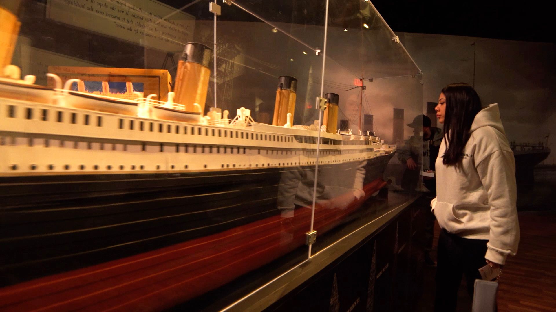 A young woman in a light gray hoodie stands before a glass display case housing a detailed white ocean liner model with an orange hull, lifeboats, and smokestacks in a dim museum.