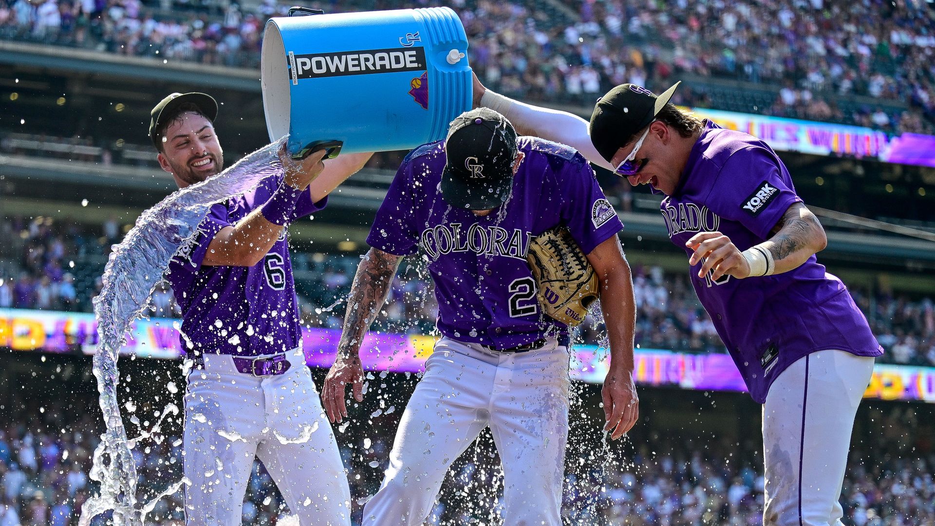 Colorado Rockies players in purple jerseys celebrate on a baseball field as a large blue Powerade cooler dumps water over them, sending spray across the white pants; crowd cheers in the stands.