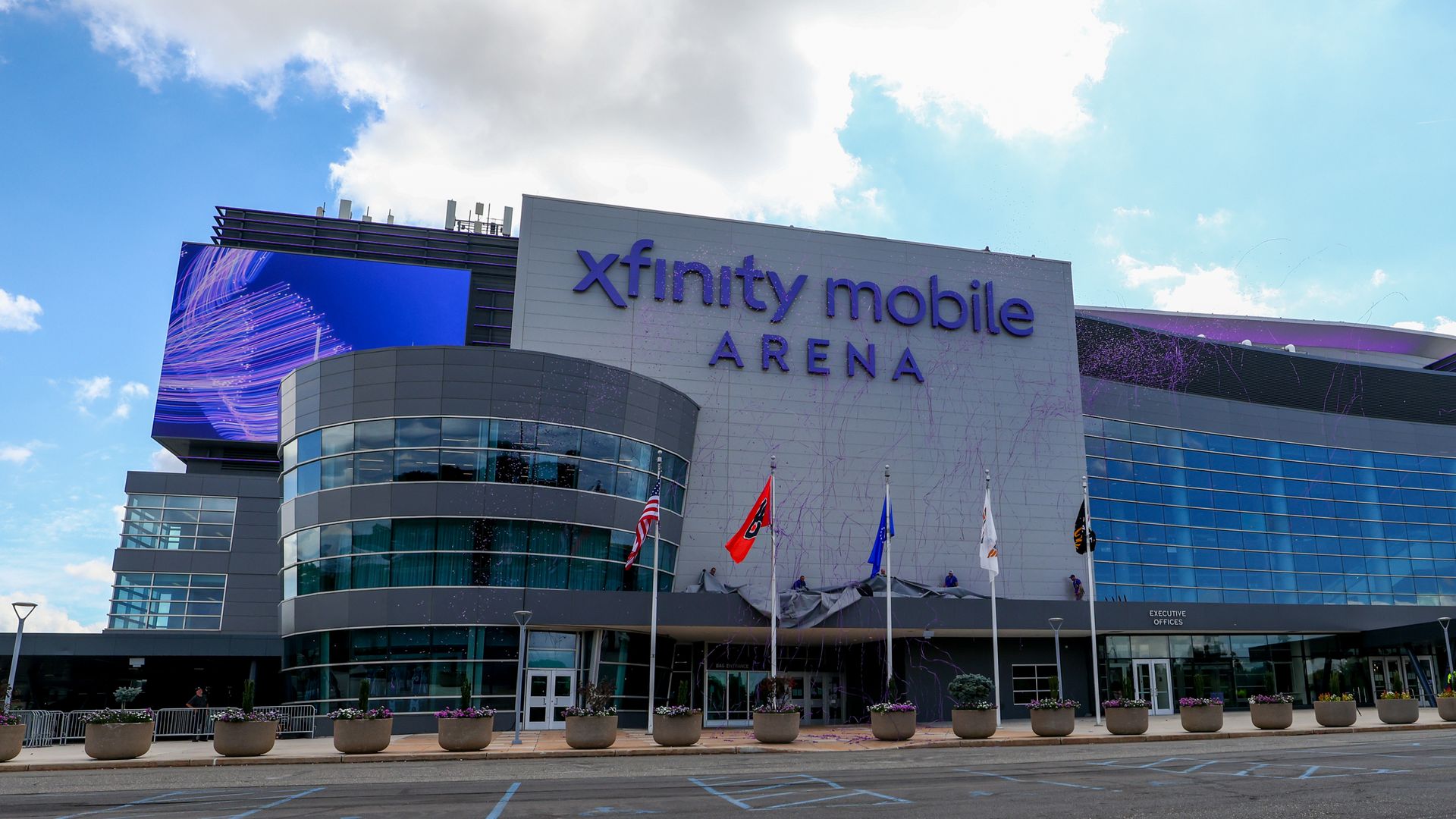 Exterior view of Xfinity Mobile Arena with modern glass facade, blue sky, scattered clouds, flags, and large digital screen on a sunny day.
