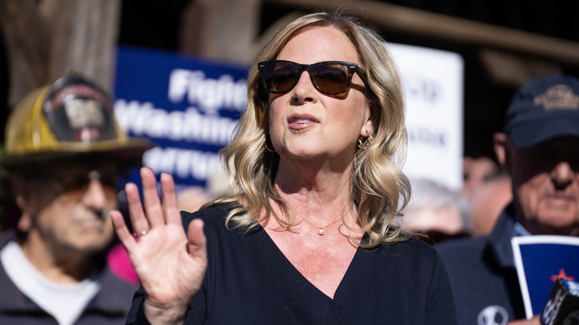 Blonde woman in a dark top wears large brown sunglasses and gold hoop earrings, raising a hand in a crowd at a rally. A blue sign and blurred protesters appear in the background.