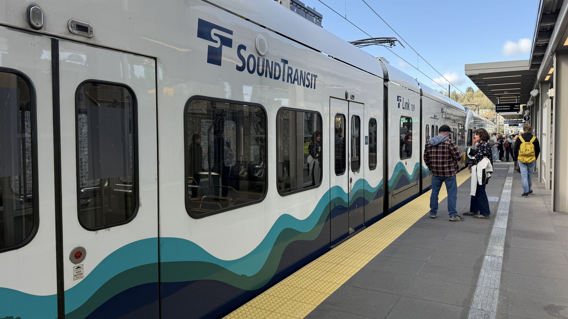 A silver light-rail train at a sunny station with blue "Sound Transit" logo and teal wave graphics along its side. Passengers wait on the platform's yellow tactile strip under overhead wires.
