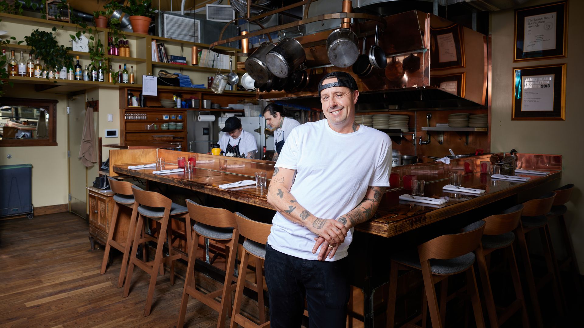 Smiling tattooed man in white shirt and cap leans on wooden counter in cozy kitchen with hanging pots, chefs cooking behind, framed awards, and potted plants on shelves.
