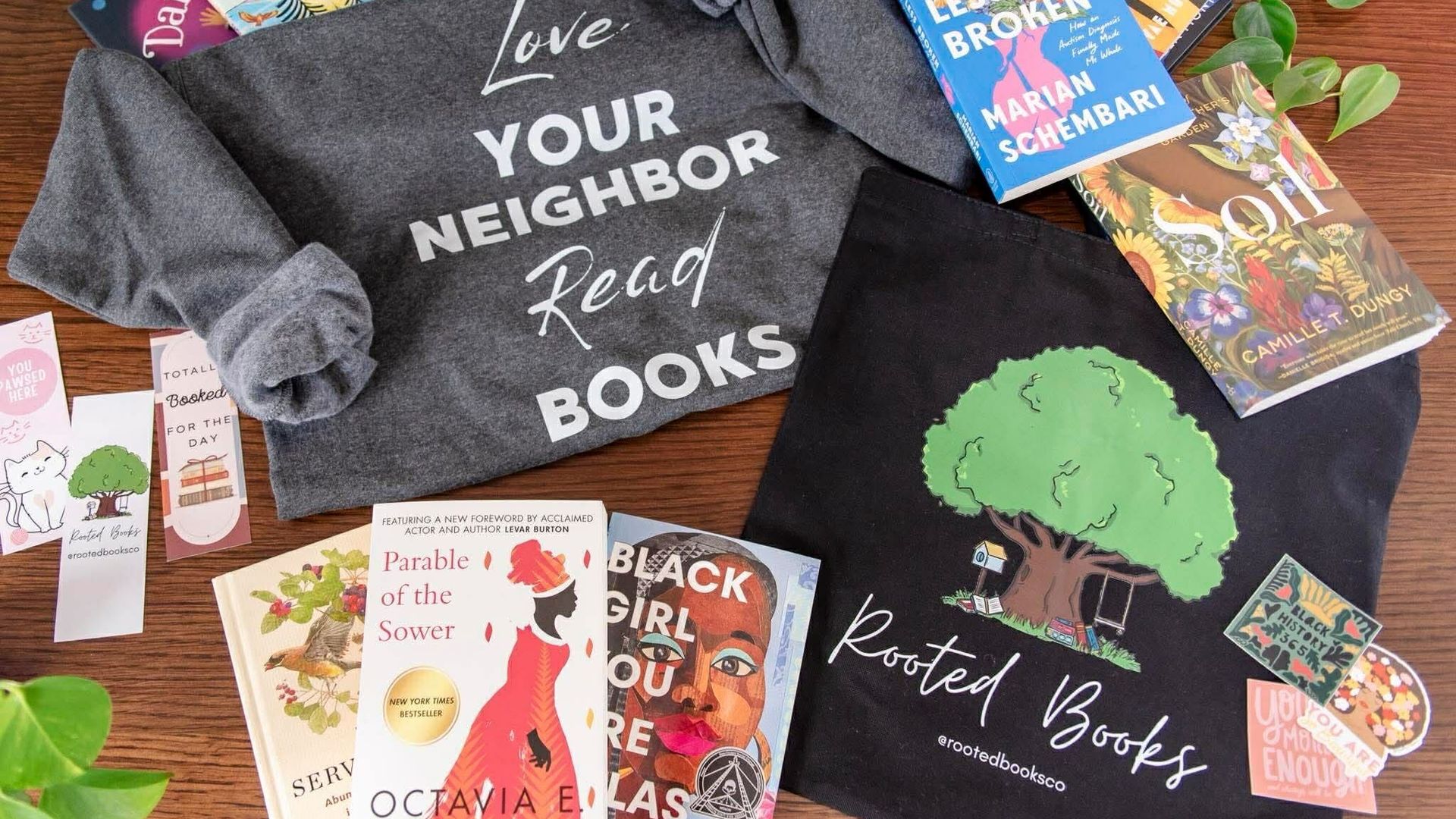 A flat lay of bookish swag: a gray hoodie reading Love Your Neighbor Read Books, a black tote with a green tree design labeled Rooted Books, book covers, bookmarks, and plants on a wooden table.