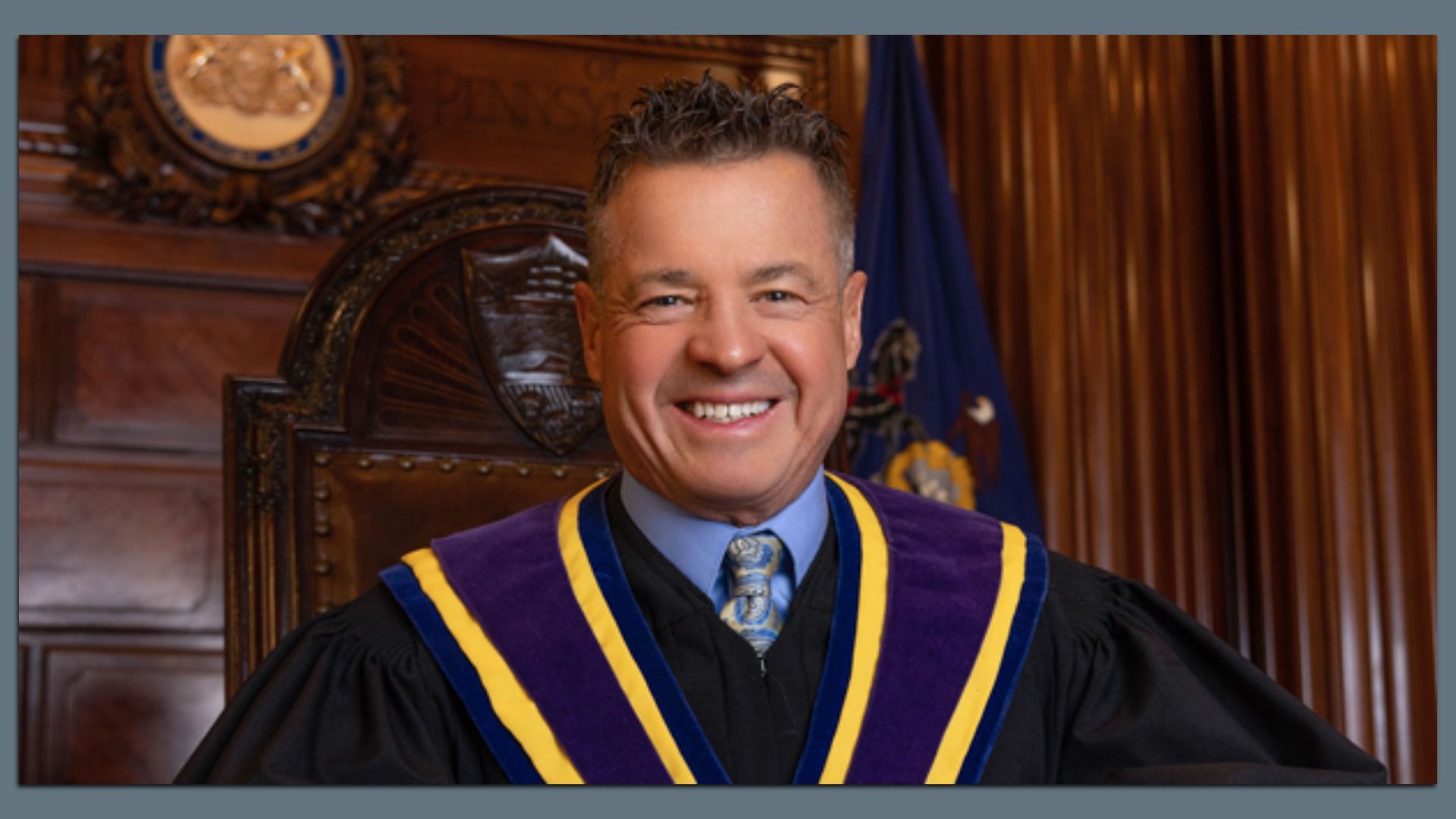 Smiling man in a black academic gown with purple and gold trim sits in a wood-paneled room, with a seal emblem and blue flag behind him.